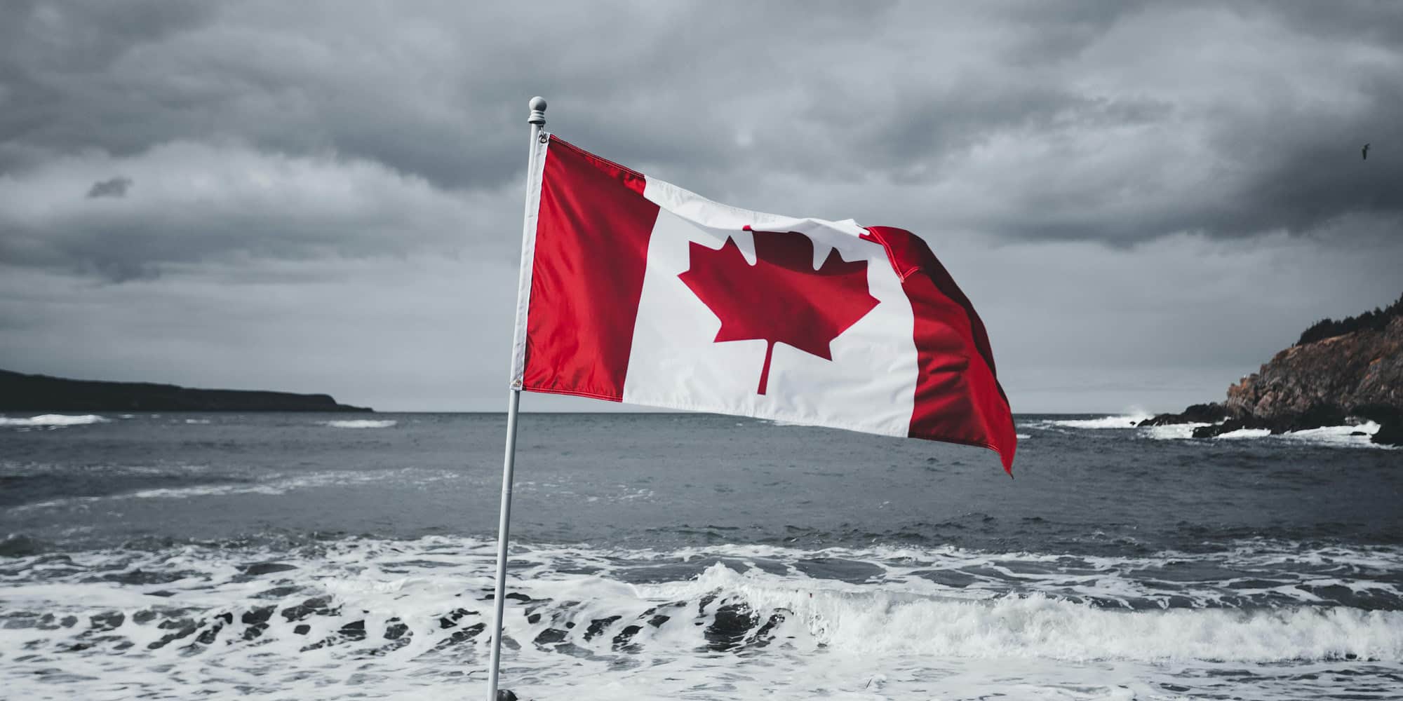 A Canadian flag with a red maple leaf waves on a pole by the ocean, with waves crashing on the shore and a cloudy, overcast sky in the background—prompting some to wonder: is one region favoured by Ottawa?.