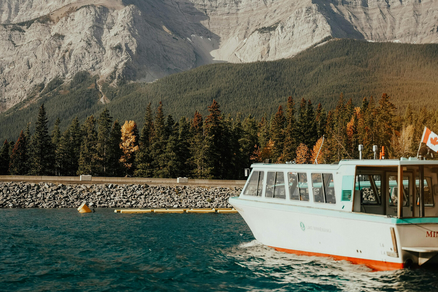 A white tour boat with a Canadian flag is docked on a turquoise lake, surrounded by evergreen trees and rocky mountains under a clear sky. Is one region favoured by Ottawa? This tranquil scene suggests nature knows no favourites.
