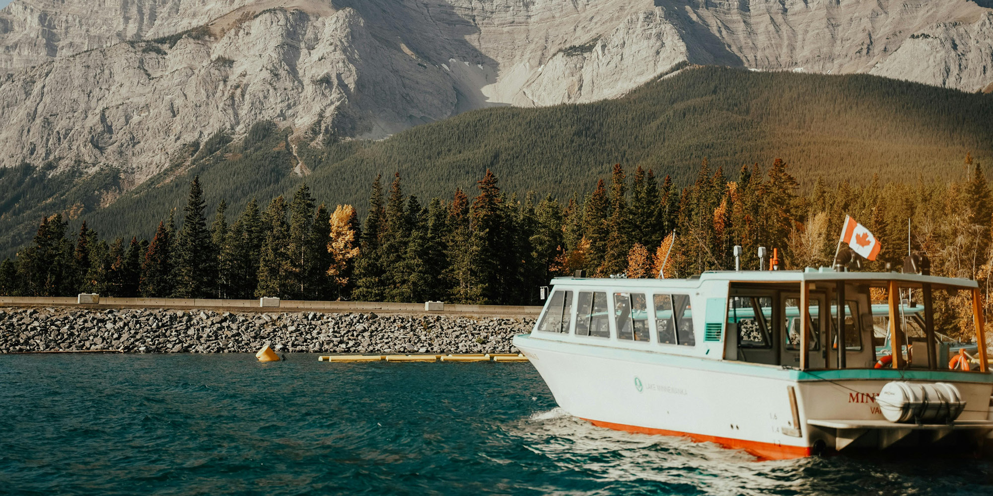 A white tour boat with a Canadian flag is docked on a turquoise lake, surrounded by evergreen trees and rocky mountains under a clear sky. Is one region favoured by Ottawa? This tranquil scene suggests nature knows no favourites.