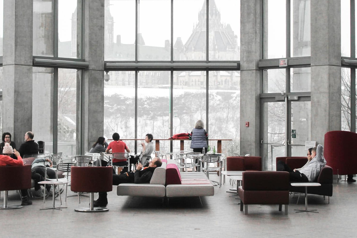 People sit and relax on modern chairs and sofas in a spacious, light-filled lounge, often exchanging Canada immigration stories, with large windows overlooking a historic-looking building and snowy landscape outside.