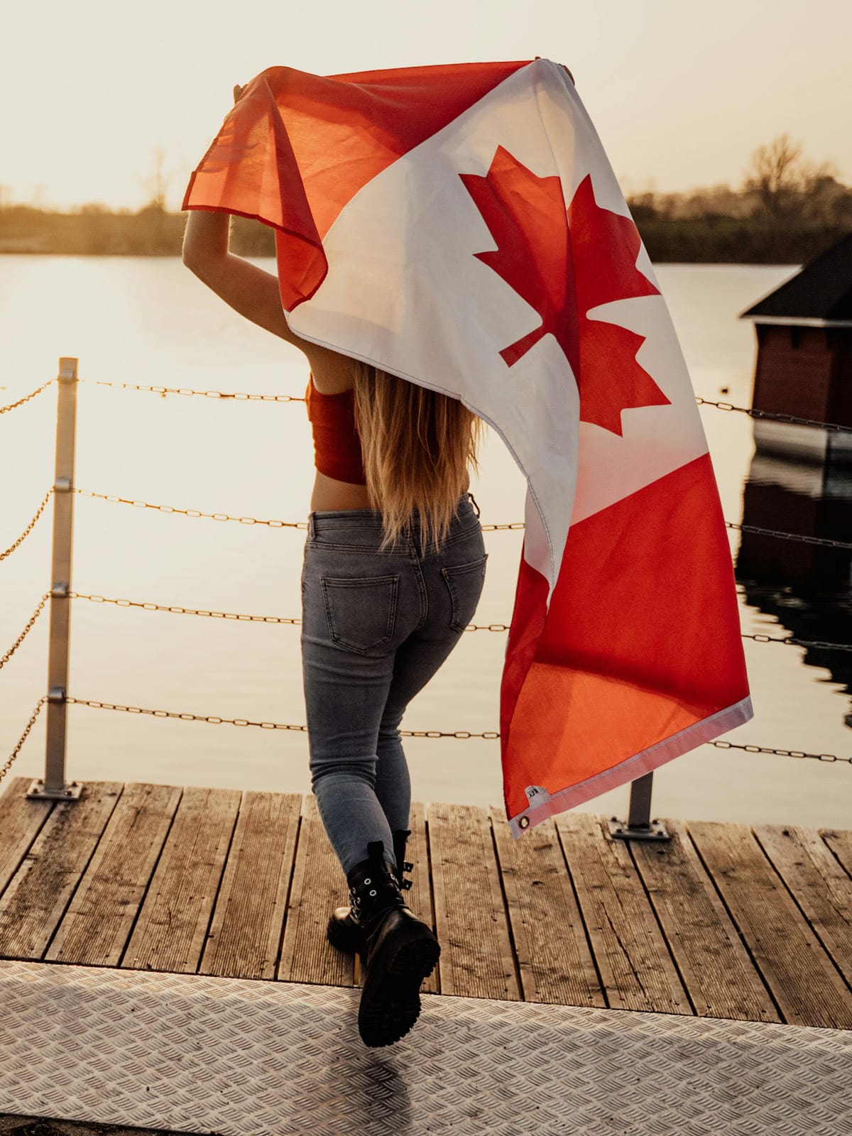 A person with long hair walks on a wooden dock by the water at sunset, draping a large Canadian flag over their shoulders. Our elbows may be up but have Canadians really changed, or do moments like this still define us?.