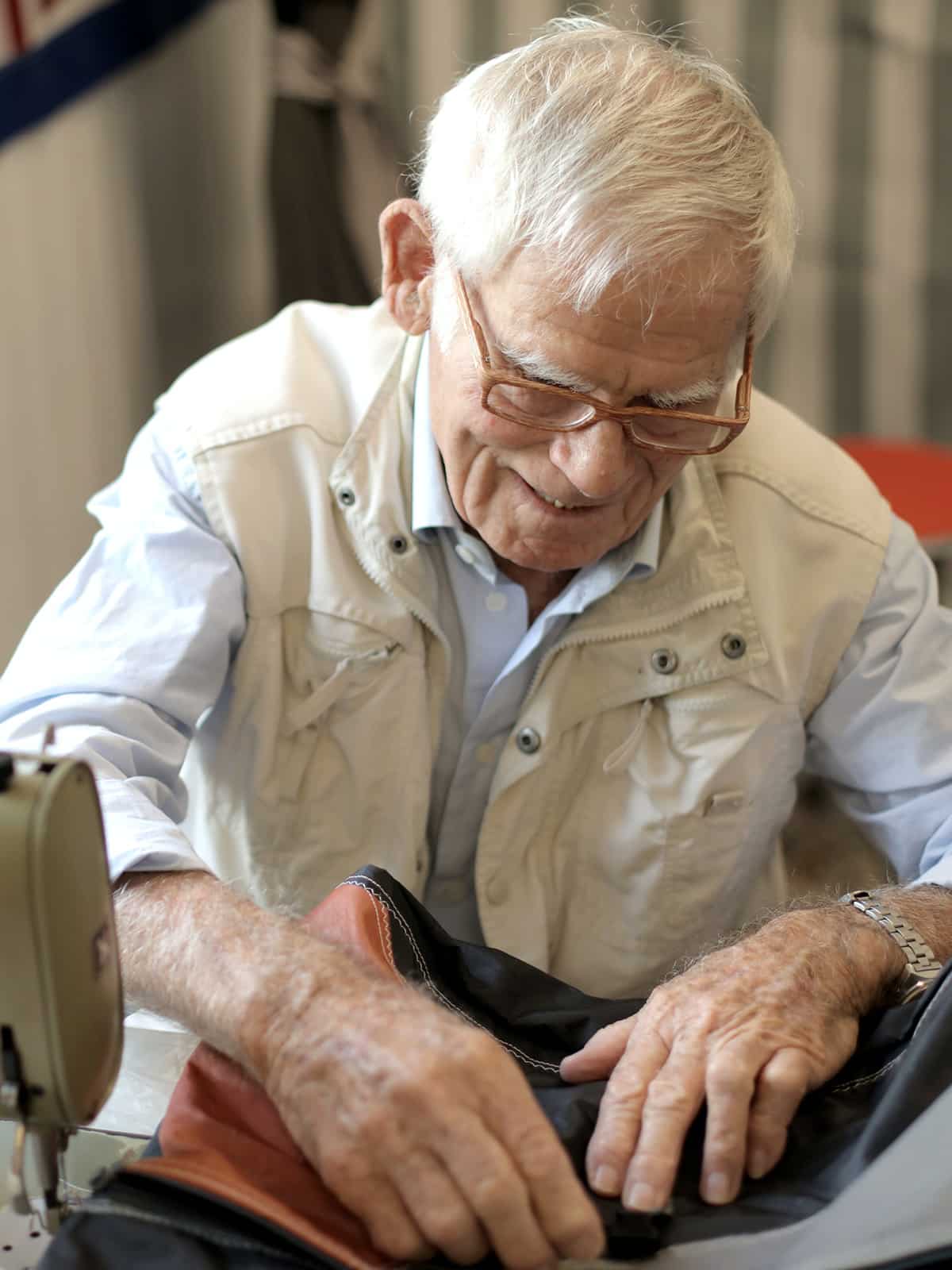 An elderly man with white hair and glasses smiles while working with fabric at a sewing machine, wearing a light blue shirt and beige vest&mdash;reminding us that our elbows may be up but have Canadians really changed?.
