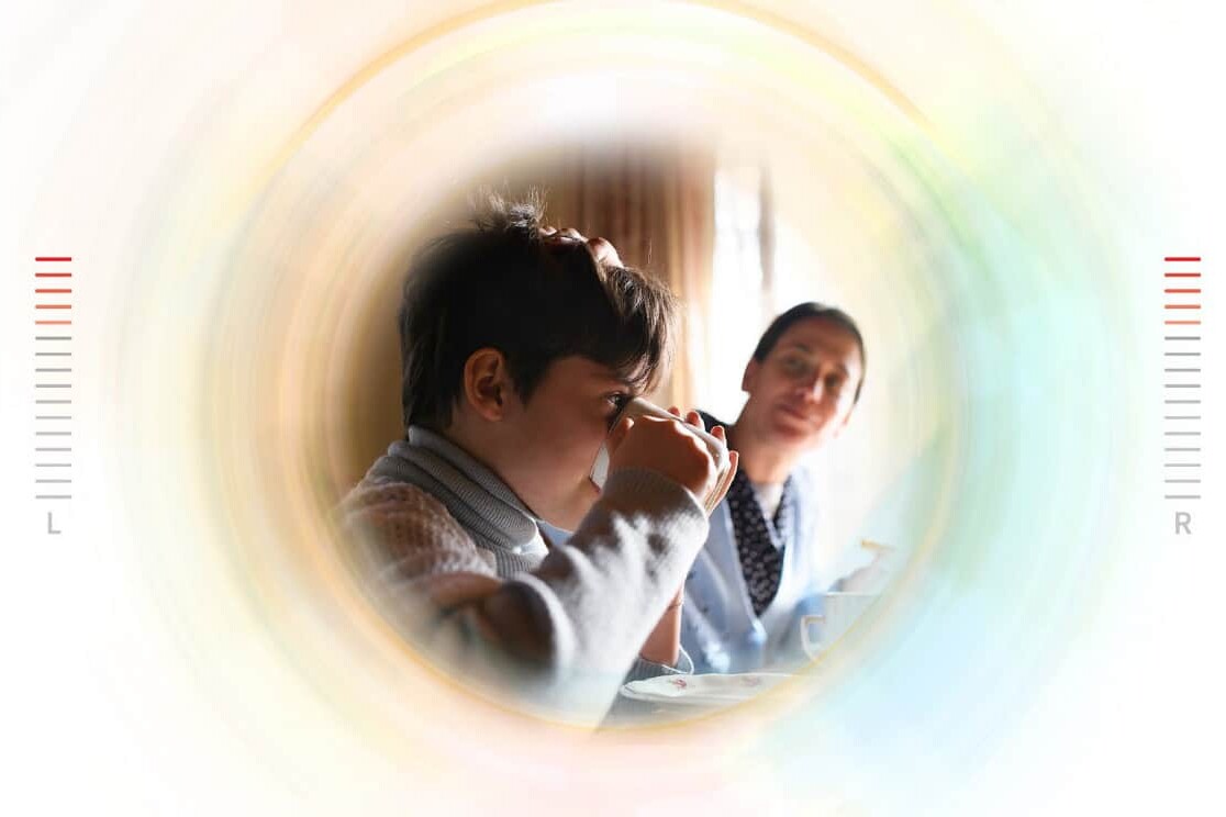 A child sits at a table drinking from a cup while an adult smiles nearby. The image, with its blurred, circular effect and L and R markers, could illustrate contexts used in developing a material deprivation index.