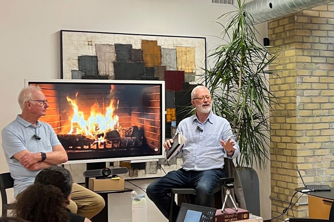 Two older men sit in front of an audience and a screen displaying a fireplace, engaged in A Social Values Journey Down Memory Lane. One gestures while speaking as the other listens, surrounded by modern décor, a large plant, and an exposed brick wall.