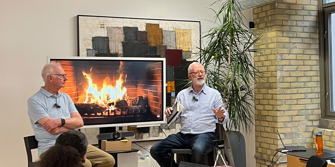Two older men sit in front of an audience and a screen displaying a fireplace, engaged in A Social Values Journey Down Memory Lane. One gestures while speaking as the other listens, surrounded by modern décor, a large plant, and an exposed brick wall.