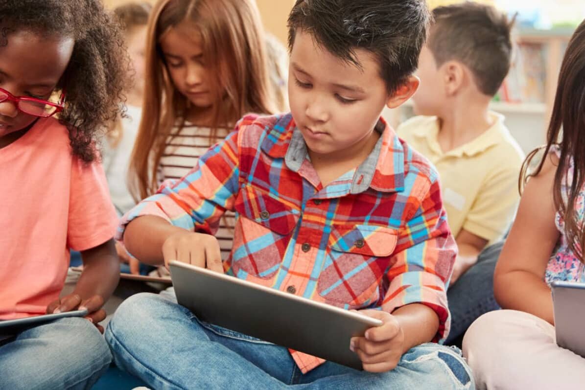 A group of young children sits on the floor in a classroom, each focused on using a tablet. The engaged students, dressed in casual clothes, reflect how Strong Schools build a Stronger Future through quality Public Education.