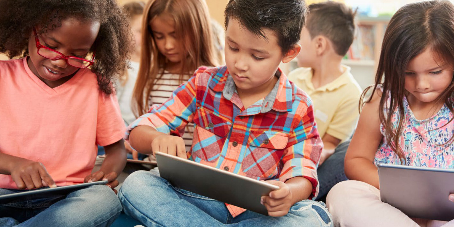 A group of young children sits on the floor in a classroom, each focused on using a tablet. The engaged students, dressed in casual clothes, reflect how Strong Schools build a Stronger Future through quality Public Education.