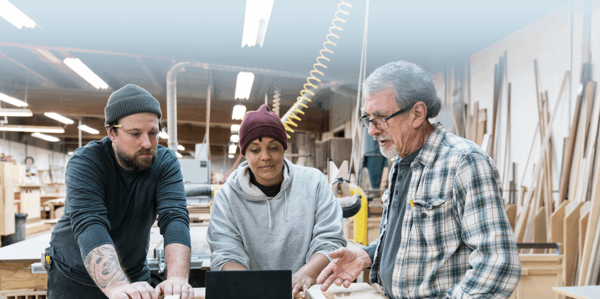 Three people stand around a workbench in a woodworking shop, looking at a tablet. As perceptions of trades training gain traction over university, they discuss their project amid tools, wooden boards, and workshop equipment.