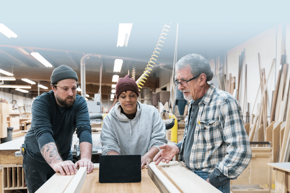 Three people stand around a workbench in a woodworking shop, looking at a tablet. As perceptions of trades training gain traction over university, they discuss their project amid tools, wooden boards, and workshop equipment.