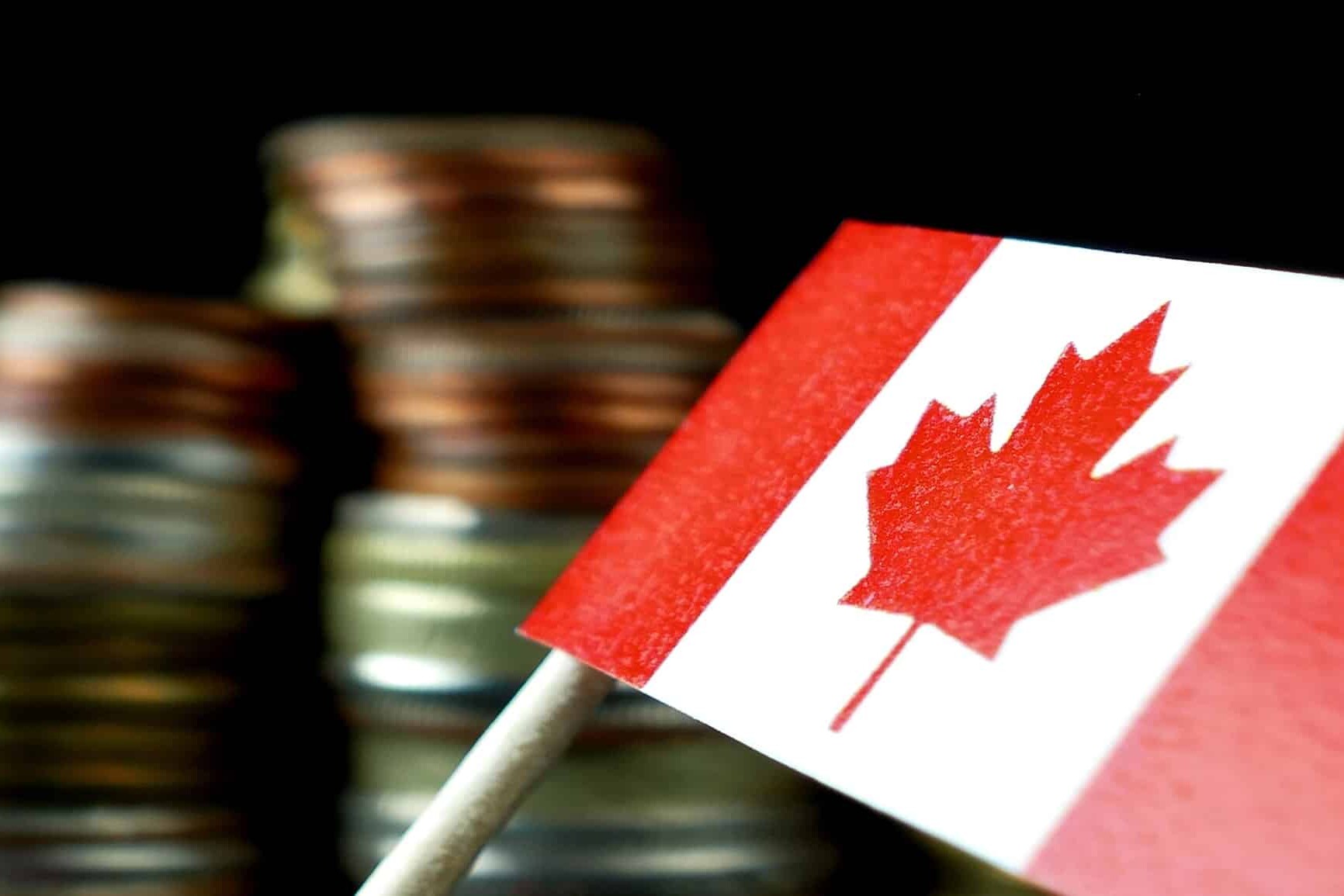 A small Canadian flag on a stick is in focus in the foreground, with blurred stacks of assorted coins—symbolizing barriers to interprovincial trade—in the background against a dark backdrop.