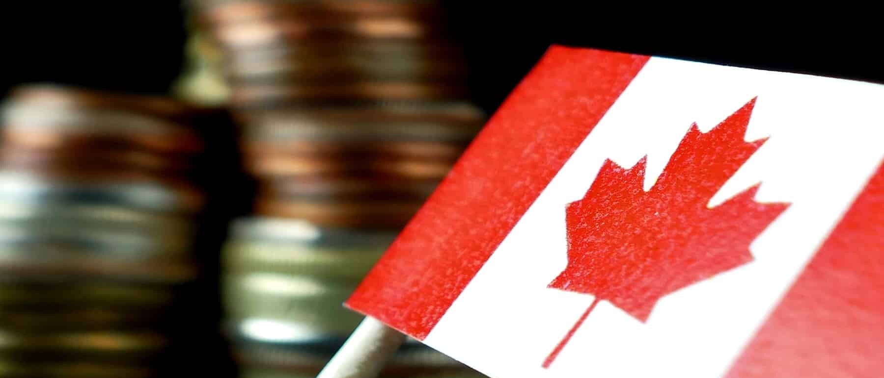 A small Canadian flag on a stick is in focus in the foreground, with blurred stacks of assorted coins—symbolizing barriers to interprovincial trade—in the background against a dark backdrop.