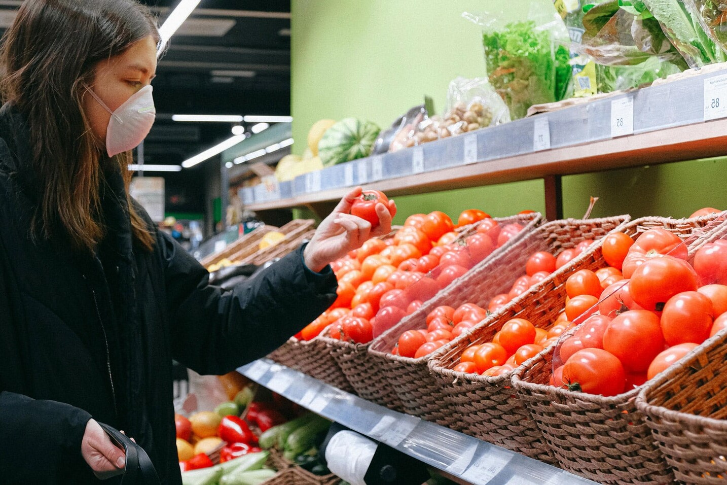 A woman wearing a face mask, reflecting on her experiences with COVID-19 and mental health, selects tomatoes from baskets filled with fresh produce in a grocery store.