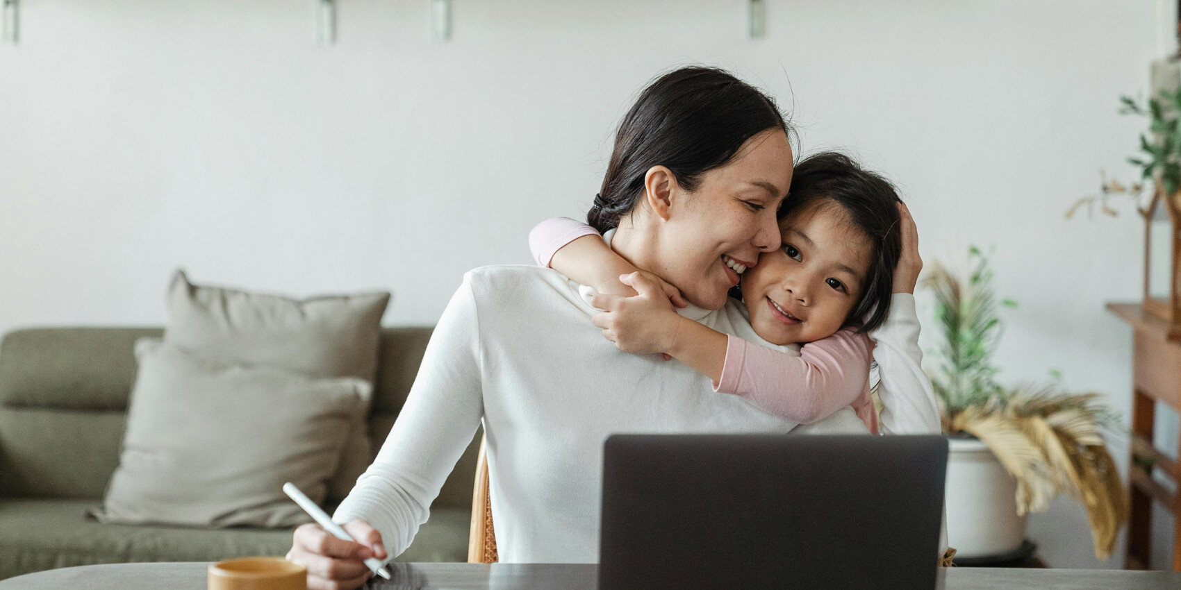 A woman sits at a table working on a tablet next to a laptop while a young girl hugs her from the side, both smiling warmly—a cozy scene reflecting the impact of having children on careers in a modern living room setting.