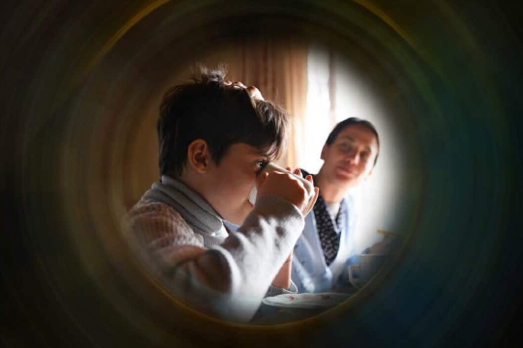 A child drinks from a cup while an adult sits nearby, watching with a gentle expression. The scene—framed softly—evokes the warmth and care that Canadian governments need as they seek new ways to measure poverty.