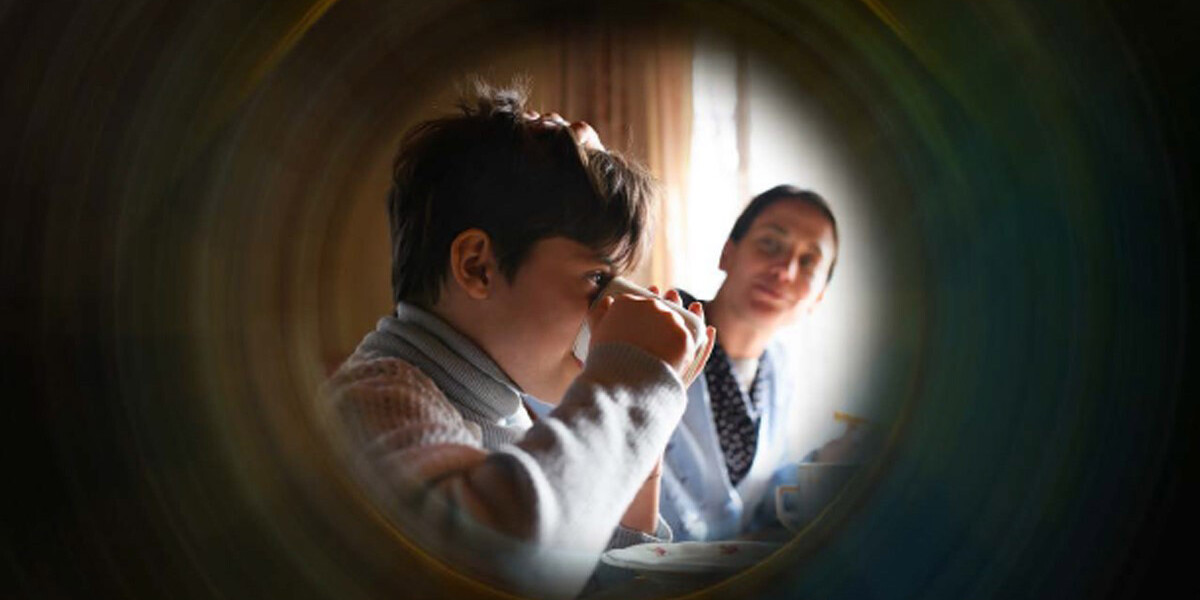 A child drinks from a cup while an adult sits nearby, watching with a gentle expression. The scene—framed softly—evokes the warmth and care that Canadian governments need as they seek new ways to measure poverty.