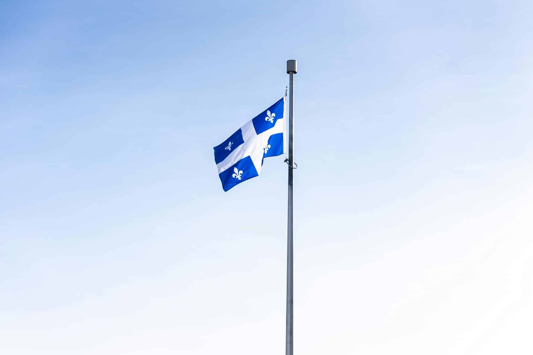 The flag of Quebec, featuring a white cross and four white fleurs-de-lis on blue, flies on a flagpole against a clear sky—a proud symbol of Canadian sovereignty in Quebec identity, culture, and language.