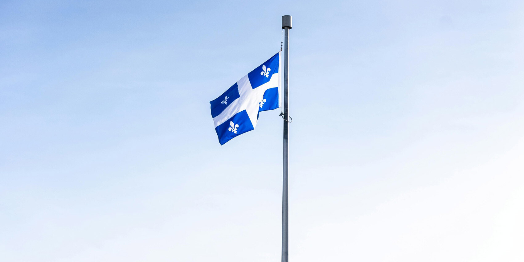 The flag of Quebec, featuring a white cross and four white fleurs-de-lis on blue, flies on a flagpole against a clear sky—a proud symbol of Canadian sovereignty in Quebec identity, culture, and language.