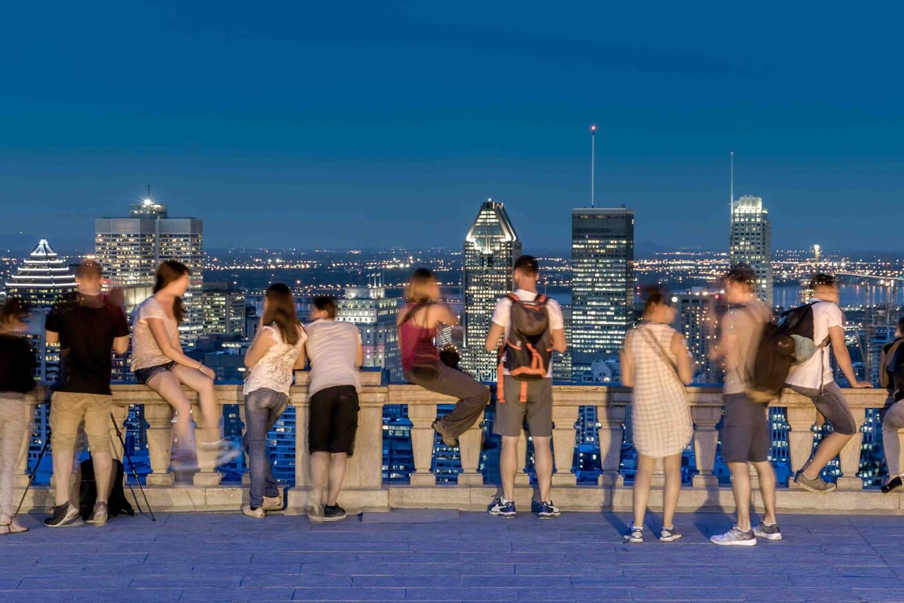 People stand and sit along a stone railing, overlooking a city skyline at dusk. The illuminated buildings and vibrant crowd reflect the rich Quebec identity, culture, and language shaped by Canadian sovereignty.