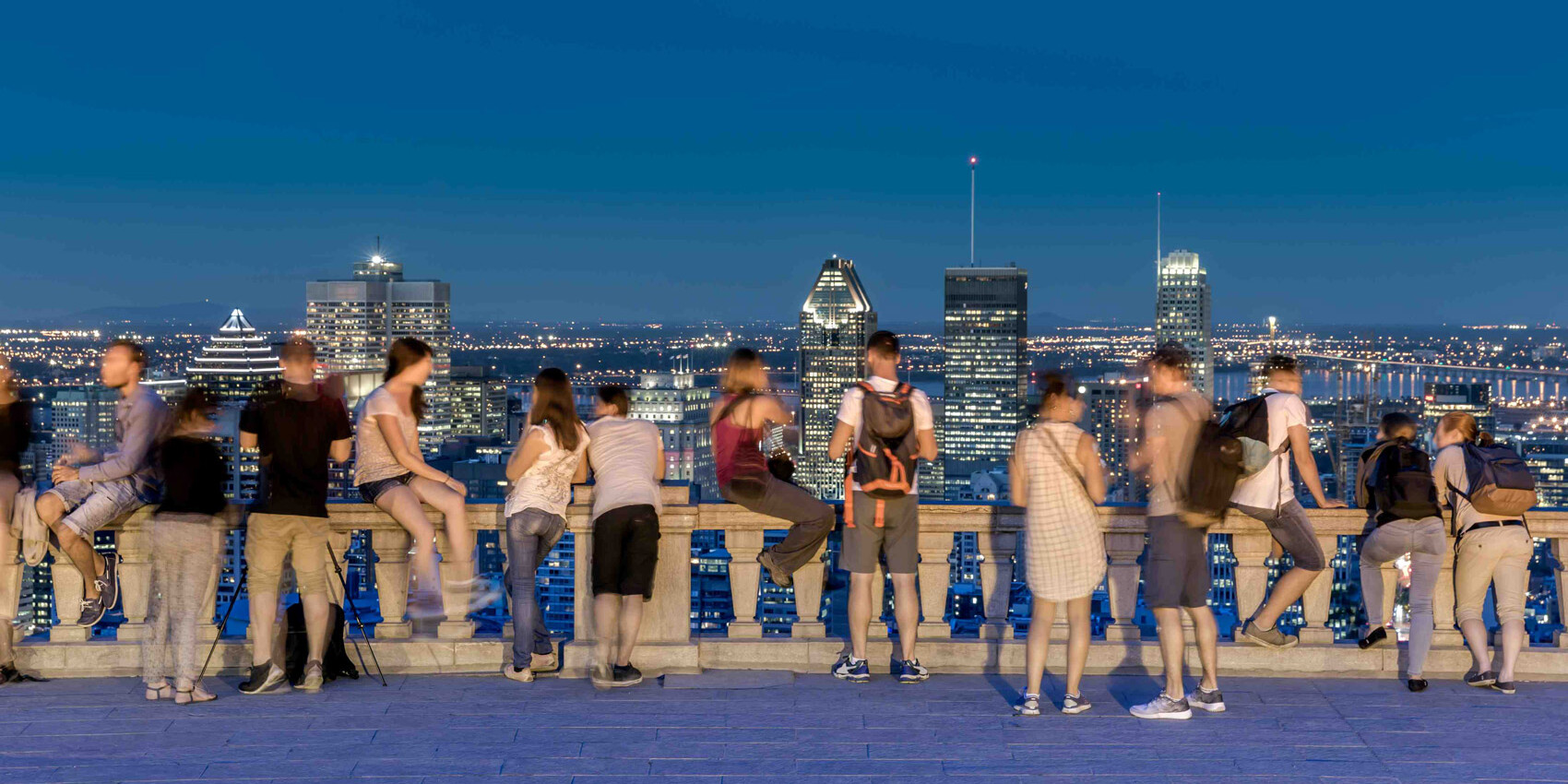 People stand and sit along a stone railing, overlooking a city skyline at dusk. The illuminated buildings and vibrant crowd reflect the rich Quebec identity, culture, and language shaped by Canadian sovereignty.