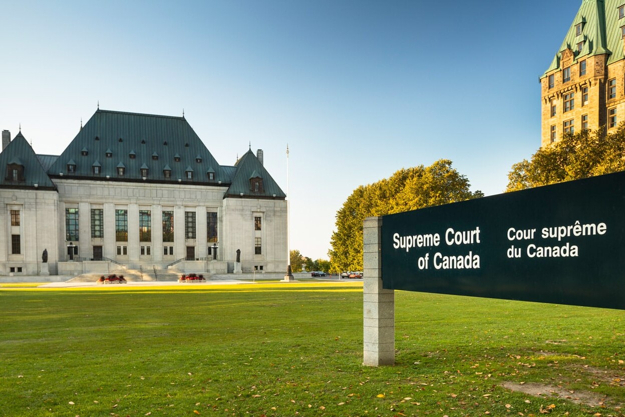 The Supreme Court of Canada building stands behind a large bilingual sign reading Supreme Court of Canada / Cour suprême du Canada, symbolizing its pivotal role in upholding the Charter of Rights, with trees and grass in the foreground under a clear sky.