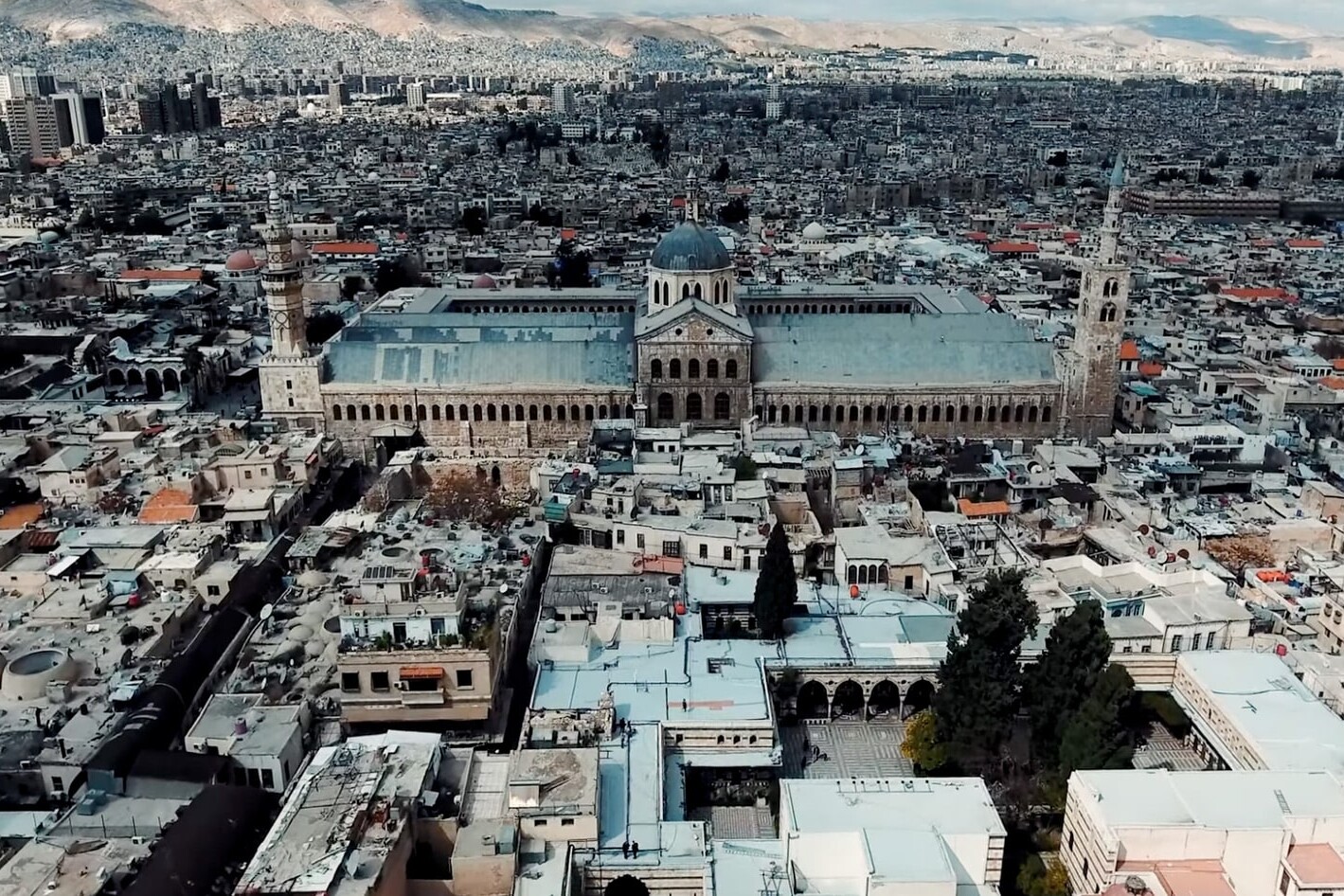 Aerial view of a large mosque with a central dome and minarets in a densely built city, surrounded by mountains. The scene highlights diversity much like a race relations Canada infographic, with historic and modern buildings side by side.
