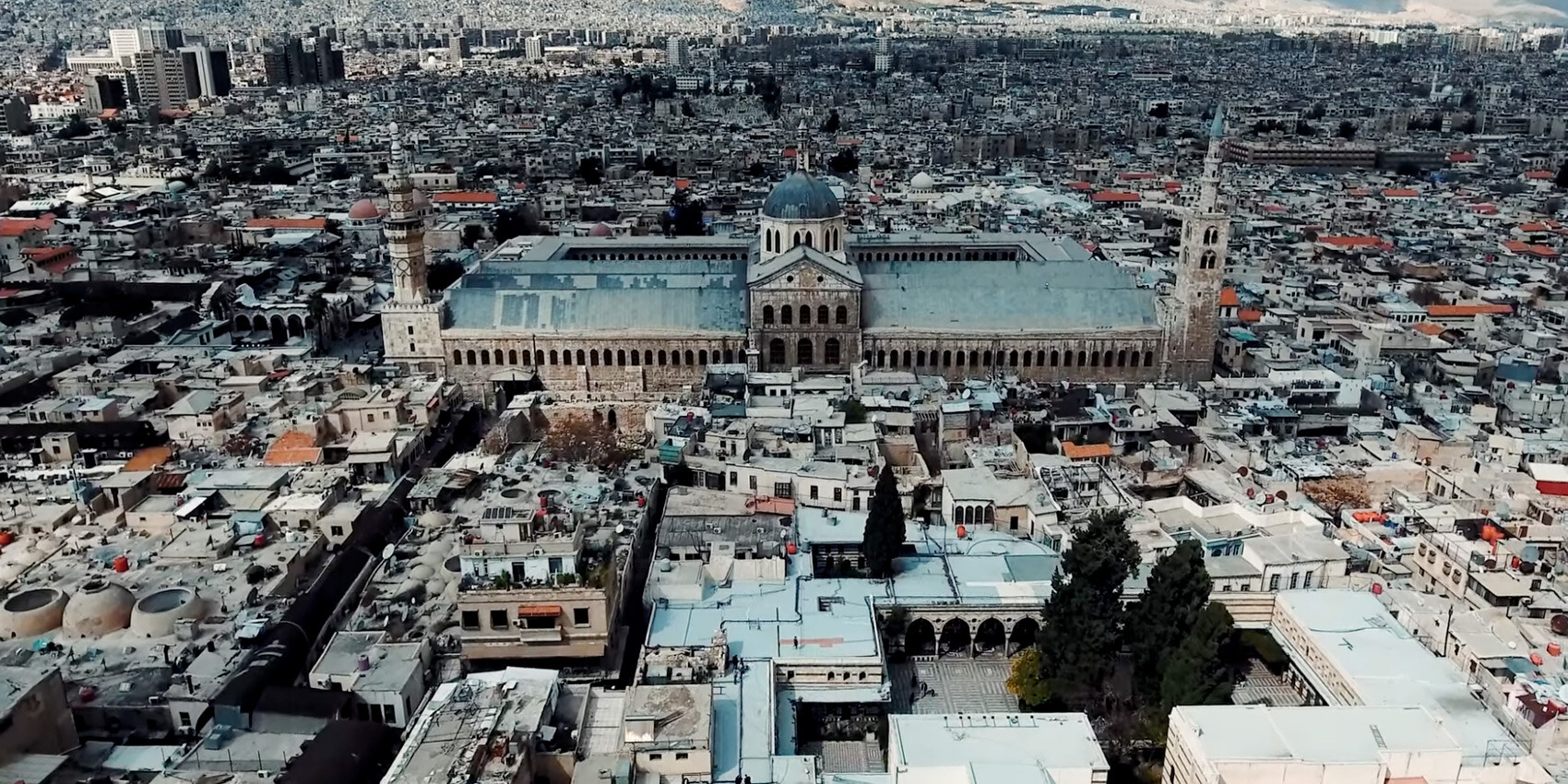 Aerial view of a large mosque with a central dome and minarets in a densely built city, surrounded by mountains. The scene highlights diversity much like a race relations Canada infographic, with historic and modern buildings side by side.