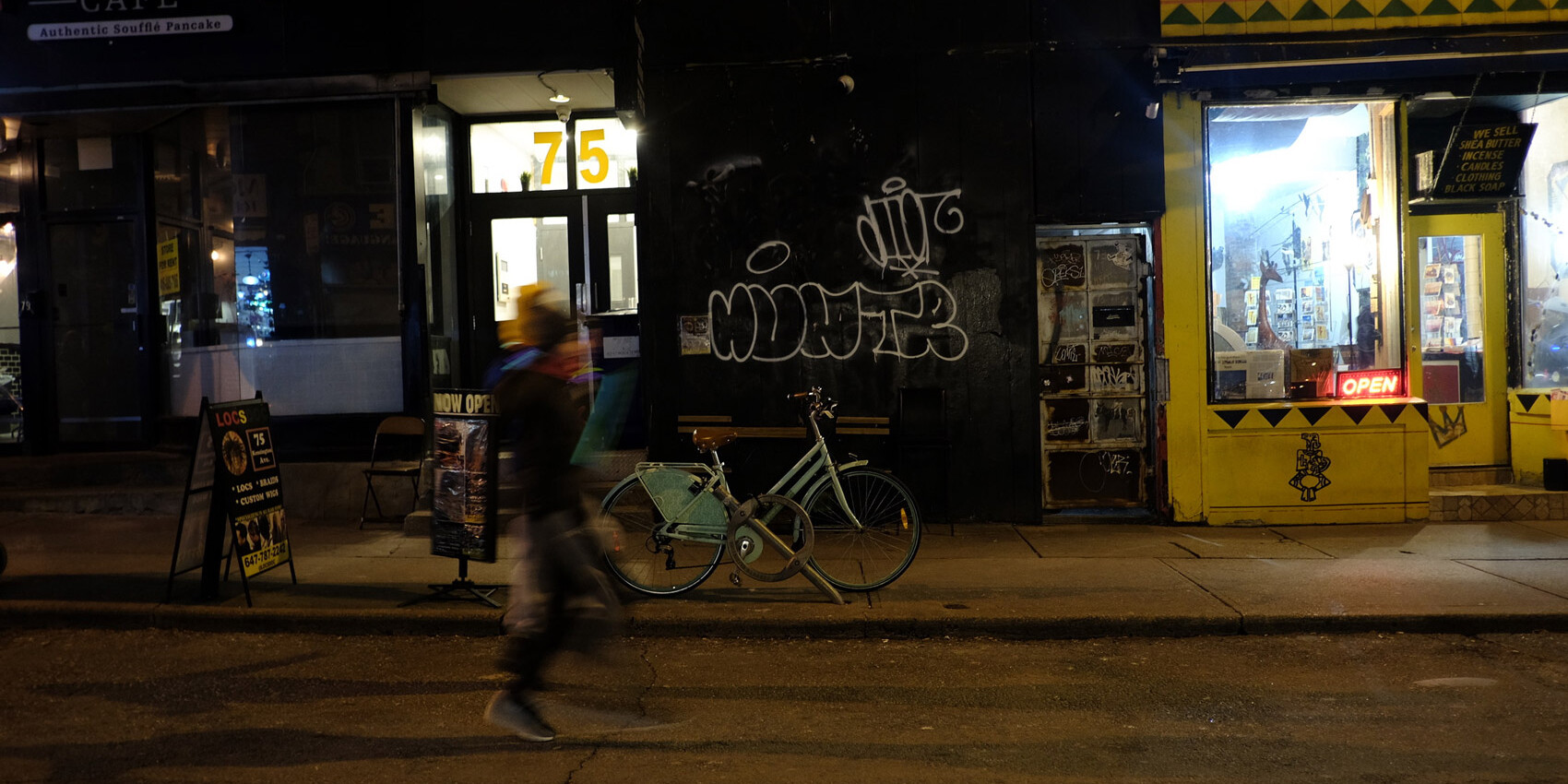 A person walks past a parked bicycle on a dimly lit city street at night. Storefronts with signs and graffiti line the background, including a yellow shop with an illuminated OPEN sign—raising the question of how to build a safer Toronto.