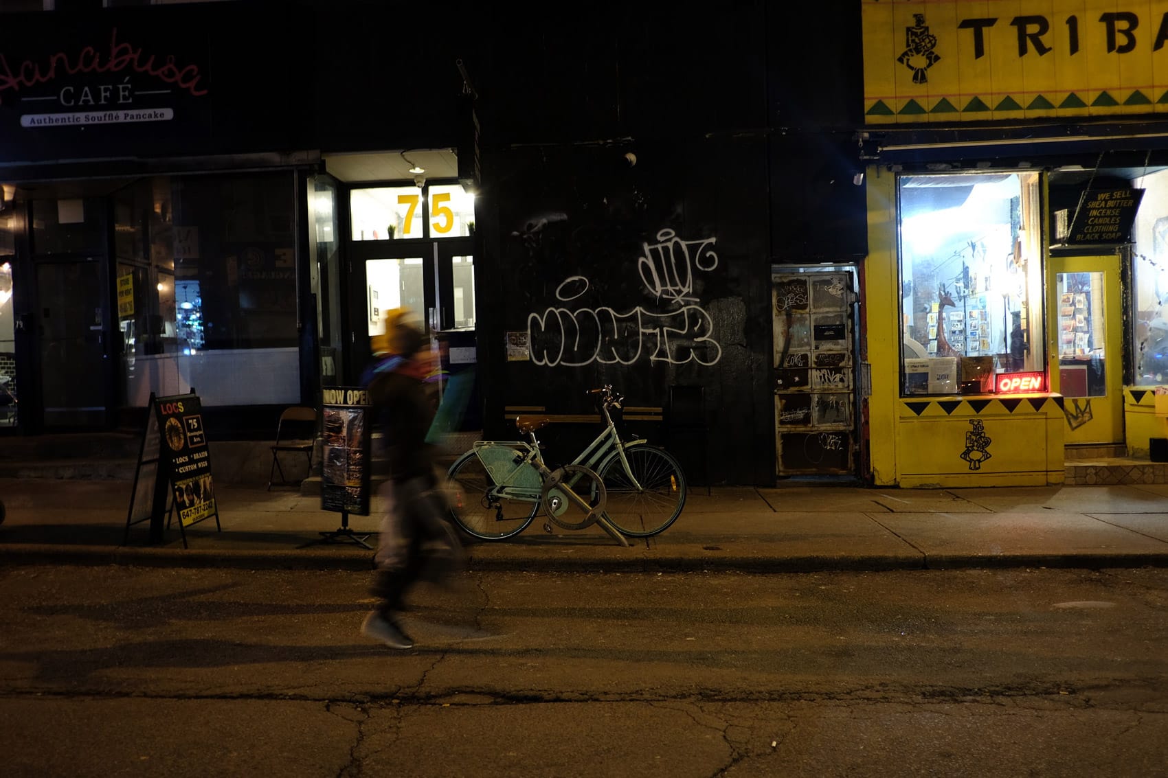A person walks past a parked bicycle on a dimly lit city street at night. Storefronts with signs and graffiti line the background, including a yellow shop with an illuminated OPEN sign—raising the question of how to build a safer Toronto.