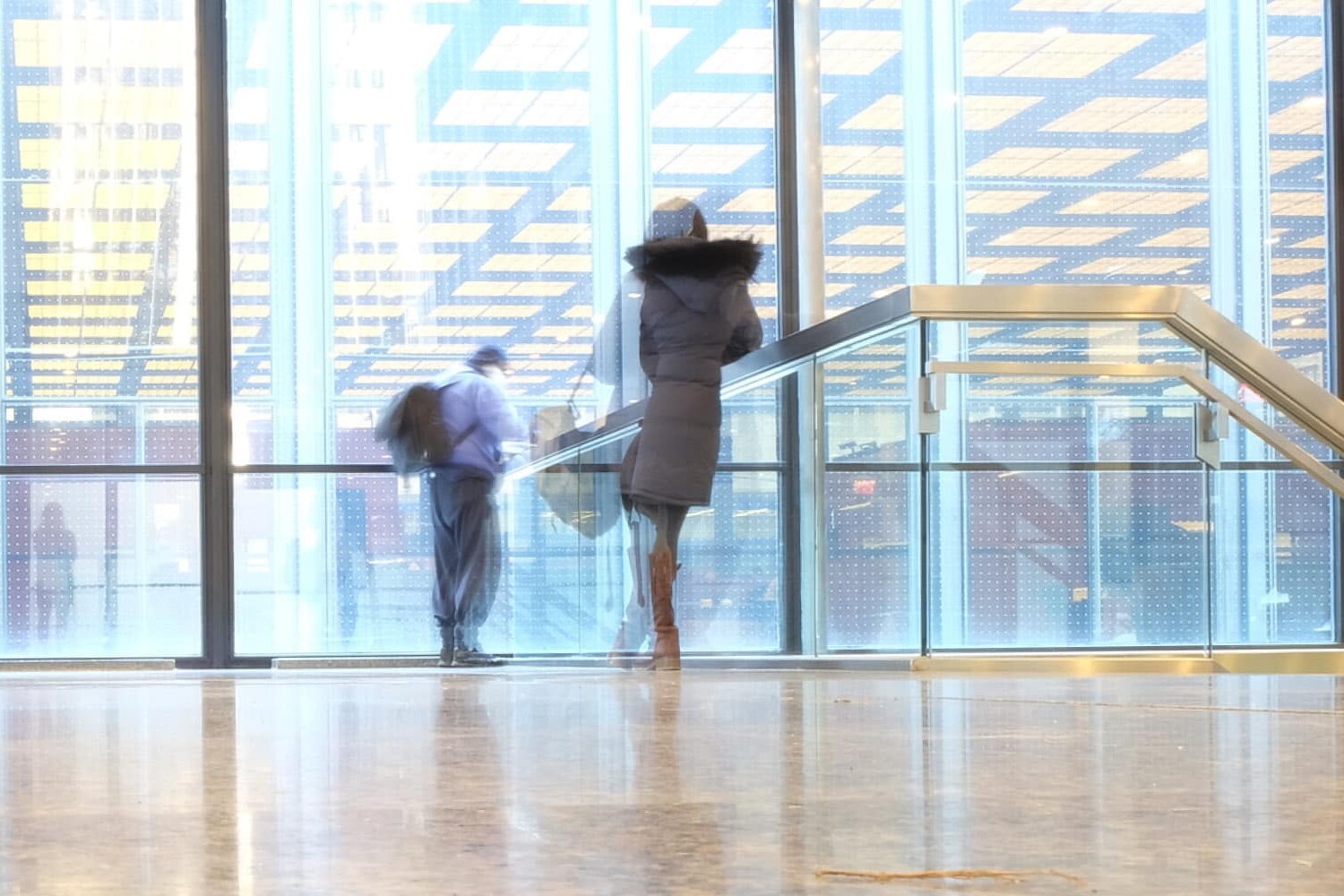 Two people, slightly blurred from motion, walk inside a modern building with large glass windows and a yellow grid ceiling. The floor is shiny, reflecting their figures—showing how coronavirus will not dent the trust Canadians have in each other.
