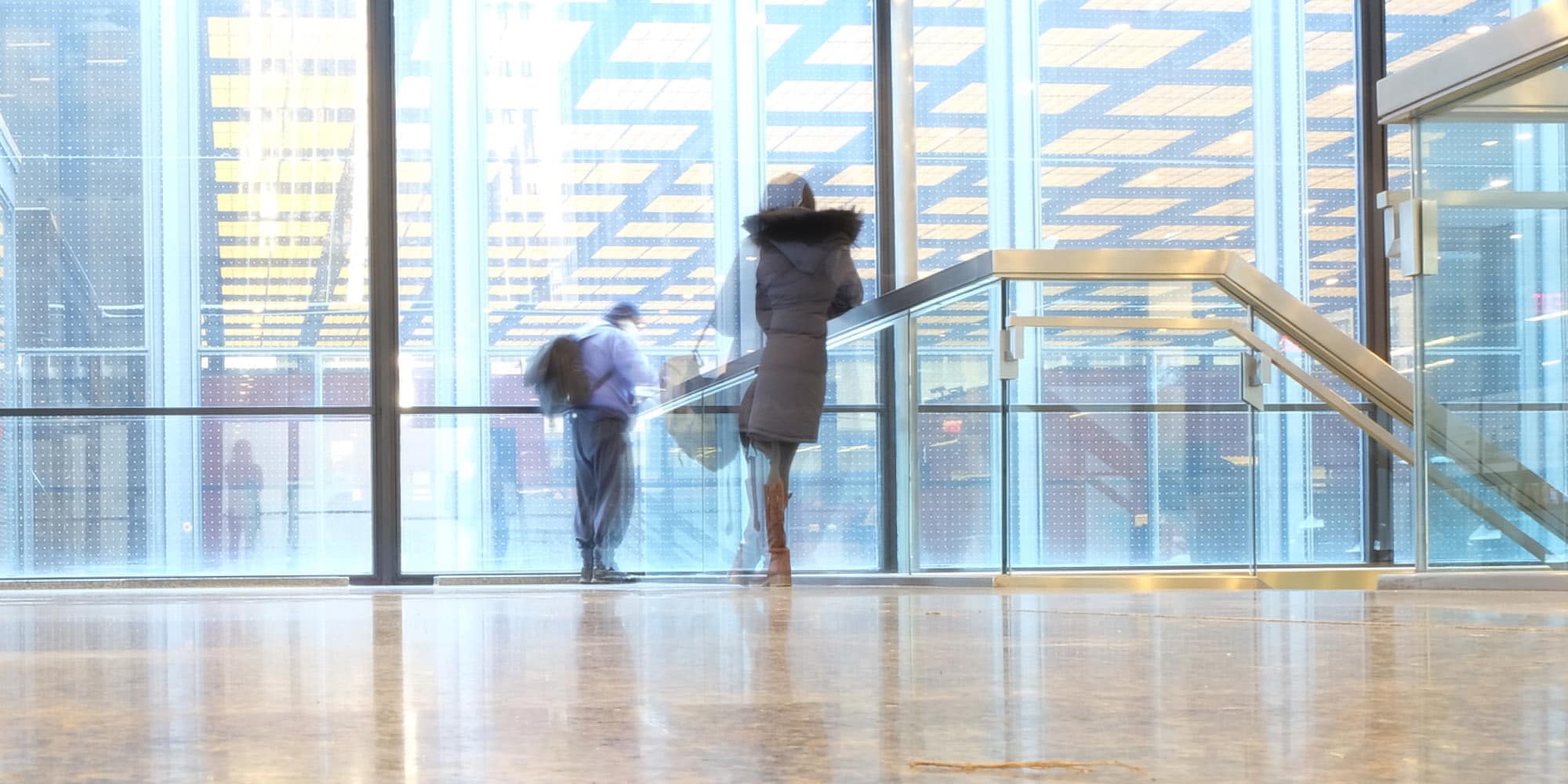 Two people, slightly blurred from motion, walk inside a modern building with large glass windows and a yellow grid ceiling. The floor is shiny, reflecting their figures—showing how coronavirus will not dent the trust Canadians have in each other.