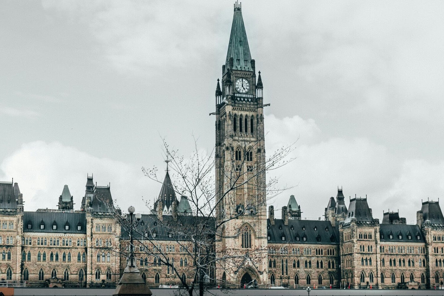 The image shows the historic Canadian Parliament building in Ottawa, featuring a central clock tower and Gothic Revival architecture under a cloudy sky, quietly prompting the question: Are Canadians finally at peace with their Constitution?.