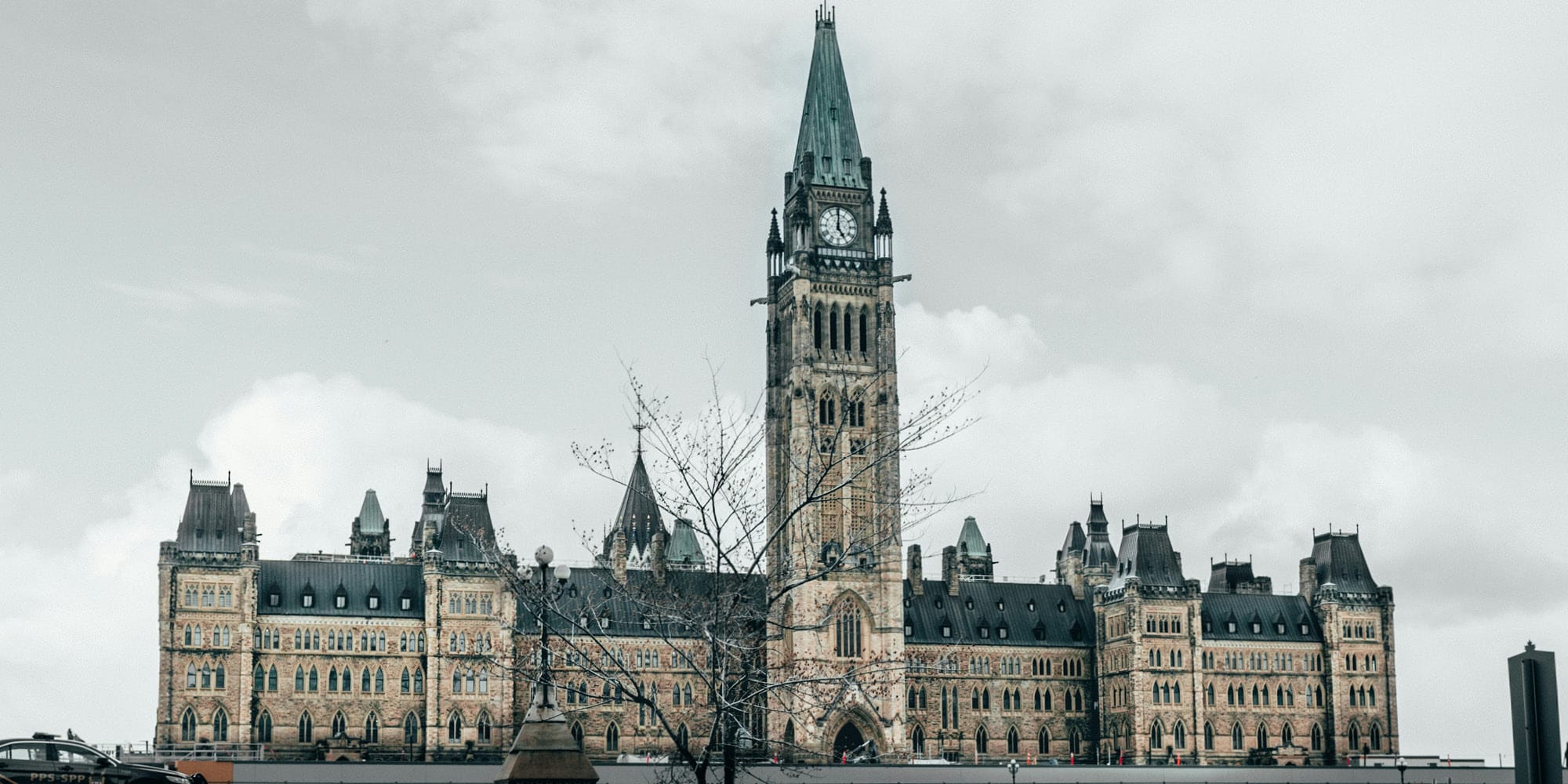 The image shows the historic Canadian Parliament building in Ottawa, featuring a central clock tower and Gothic Revival architecture under a cloudy sky, quietly prompting the question: Are Canadians finally at peace with their Constitution?.