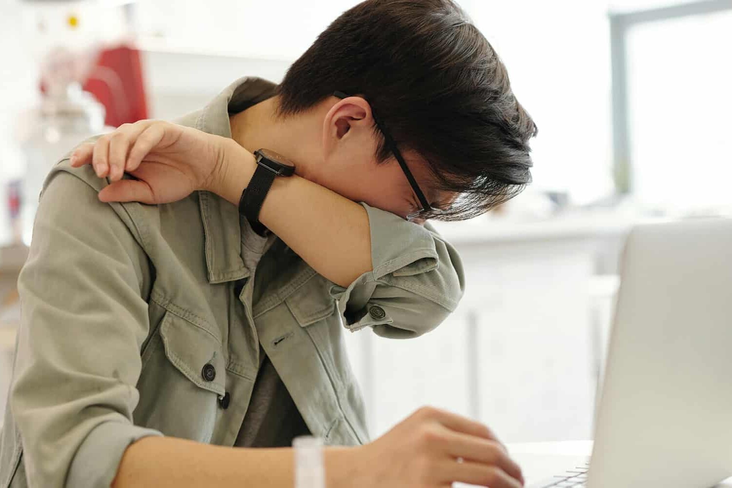 A person sitting at a table with a laptop, covering their face with their arm as if sneezing or feeling unwell, in a bright indoor setting—here’s how we can stop sick people from going into work.