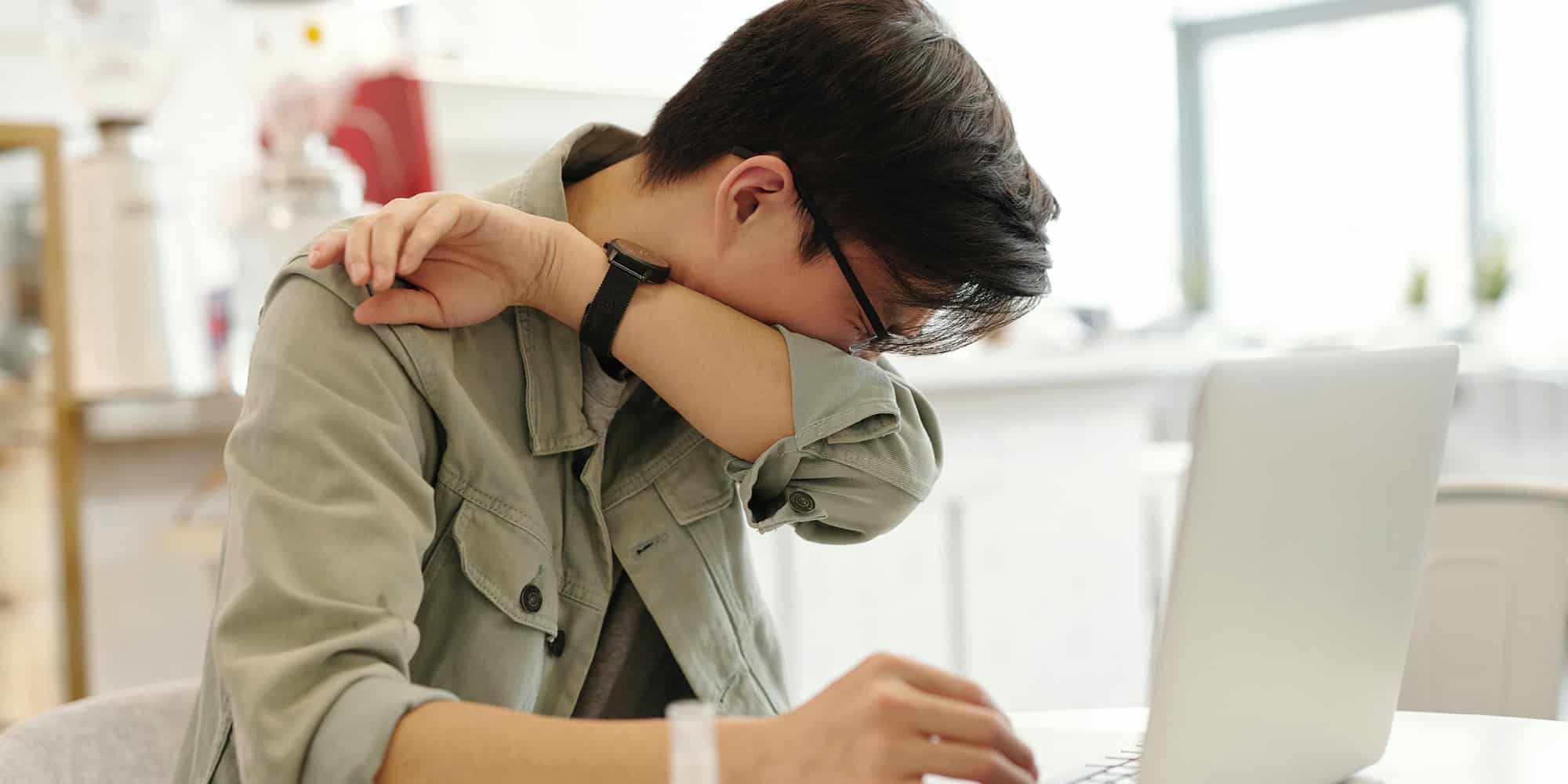A person sitting at a table with a laptop, covering their face with their arm as if sneezing or feeling unwell, in a bright indoor setting—here’s how we can stop sick people from going into work.