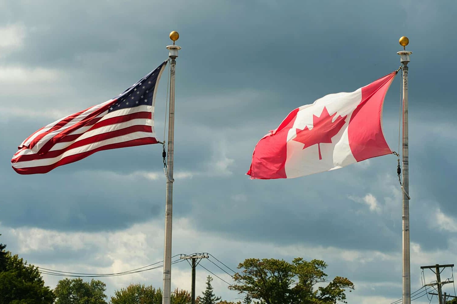 Two flags, the United States on the left and Canada on the right, wave on tall flagpoles against a cloudy sky with trees and utility poles in the background, symbolizing the continuing cultural divergence of Canada and the United States.
