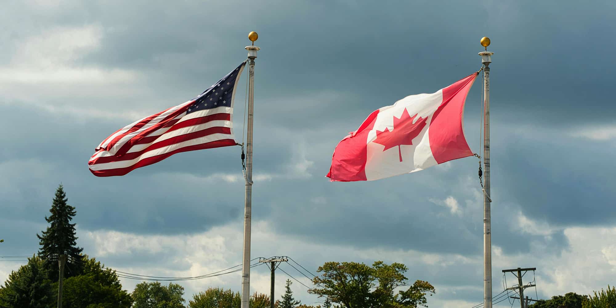 Two flags, the United States on the left and Canada on the right, wave on tall flagpoles against a cloudy sky with trees and utility poles in the background, symbolizing the continuing cultural divergence of Canada and the United States.