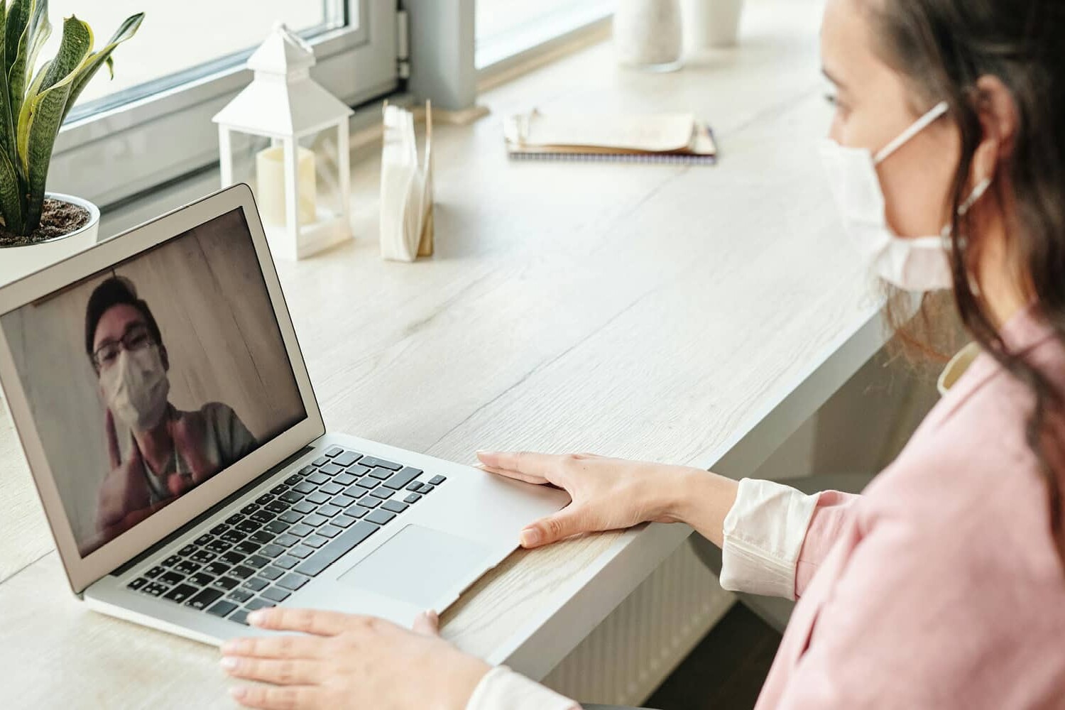 A woman wearing a face mask sits at a desk by a window, video chatting with another masked person on her laptop. A potted plant and notebook are also on the desk, as public opinion in Canada has been shifting but not because of the pandemic.