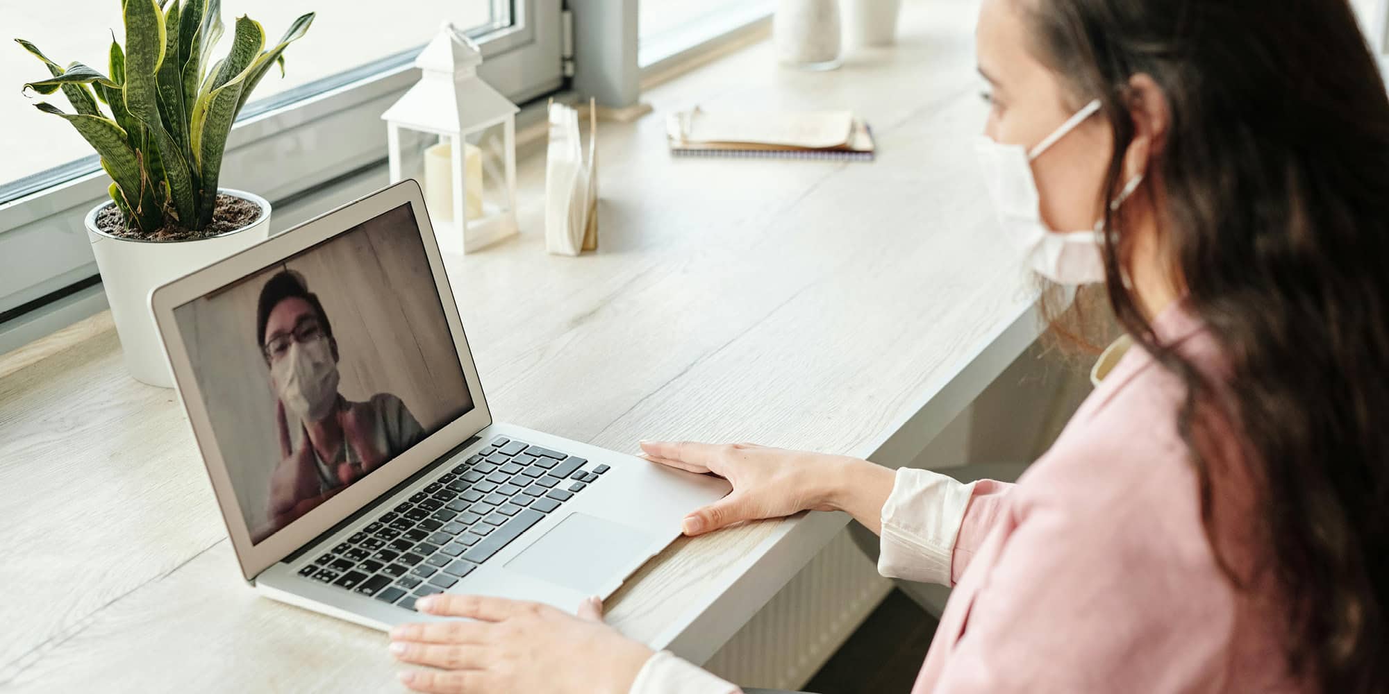 A woman wearing a face mask sits at a desk by a window, video chatting with another masked person on her laptop. A potted plant and notebook are also on the desk, as public opinion in Canada has been shifting but not because of the pandemic.