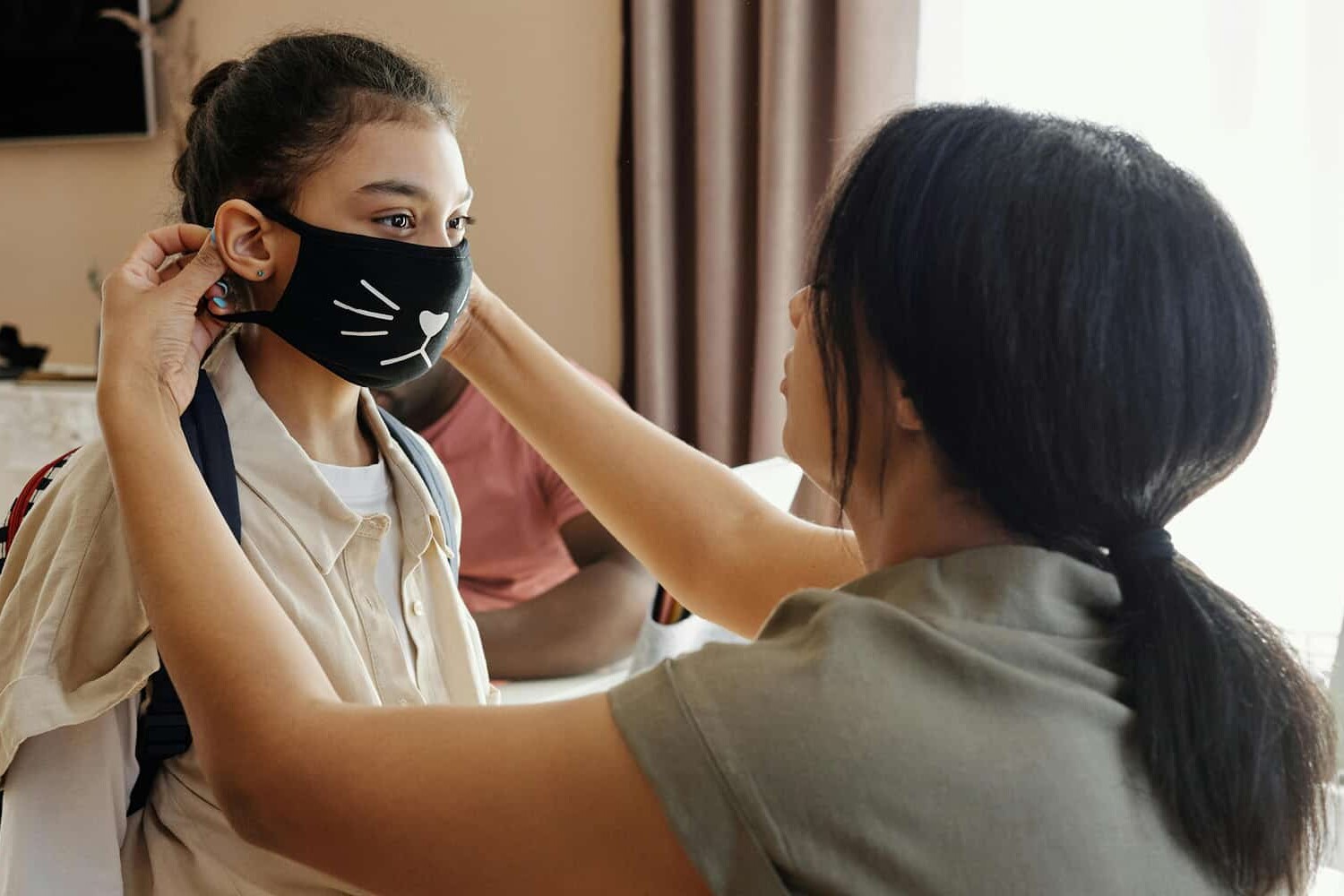 An adult helps a young girl adjust her black face mask with a cat design. The girl, wearing a backpack and school uniform, stands indoors in a well-lit room—a scene reflecting how public opinion in Canada has been shifting but not because of the pandemic.