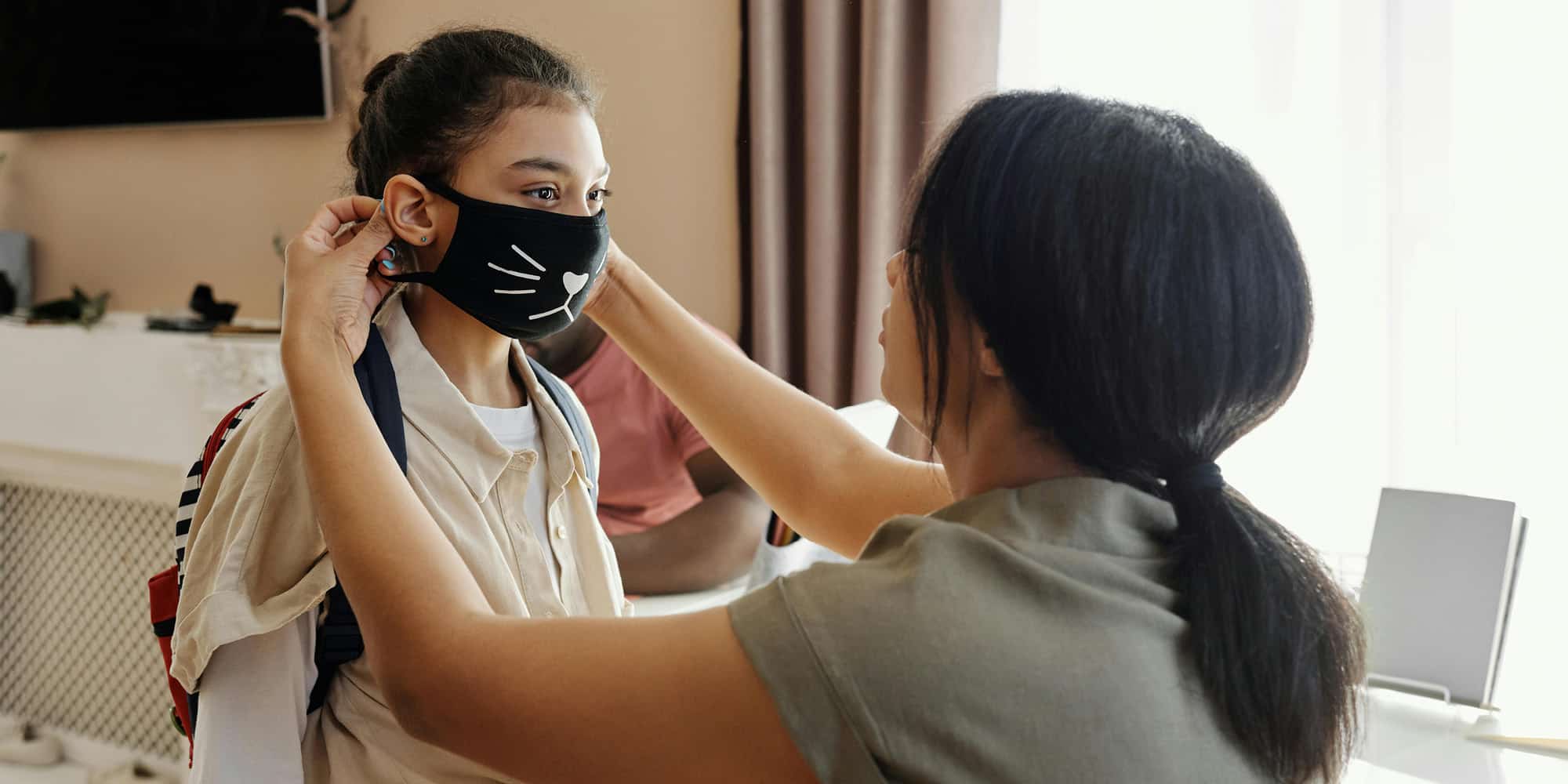 An adult helps a young girl adjust her black face mask with a cat design. The girl, wearing a backpack and school uniform, stands indoors in a well-lit room—a scene reflecting how public opinion in Canada has been shifting but not because of the pandemic.
