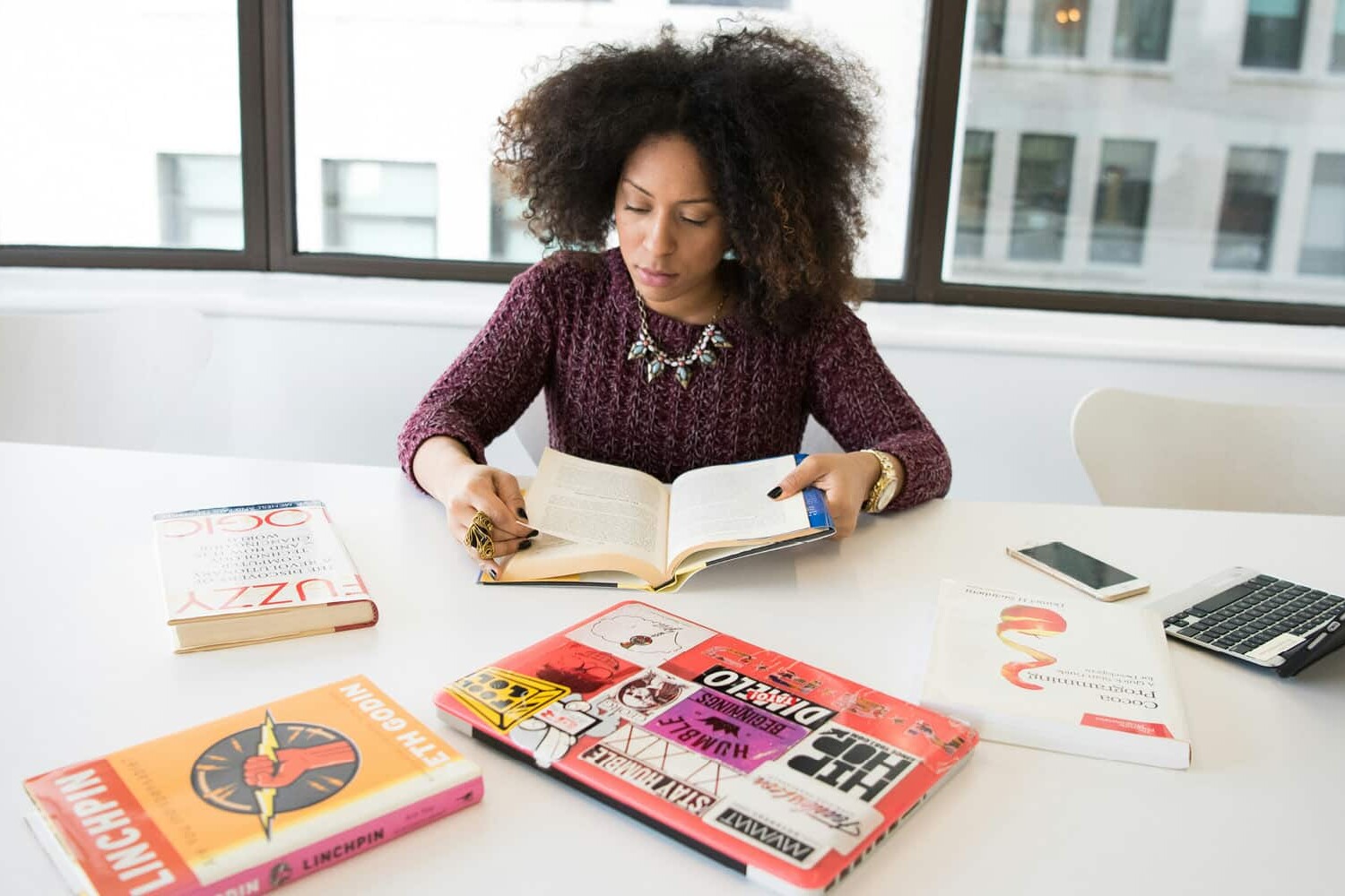 A woman with curly hair sits at a white table reading a book about the reasons why women and men are self-employed. Several books, a smartphone, and a laptop covered in stickers are on the table. Large windows reveal buildings outside.
