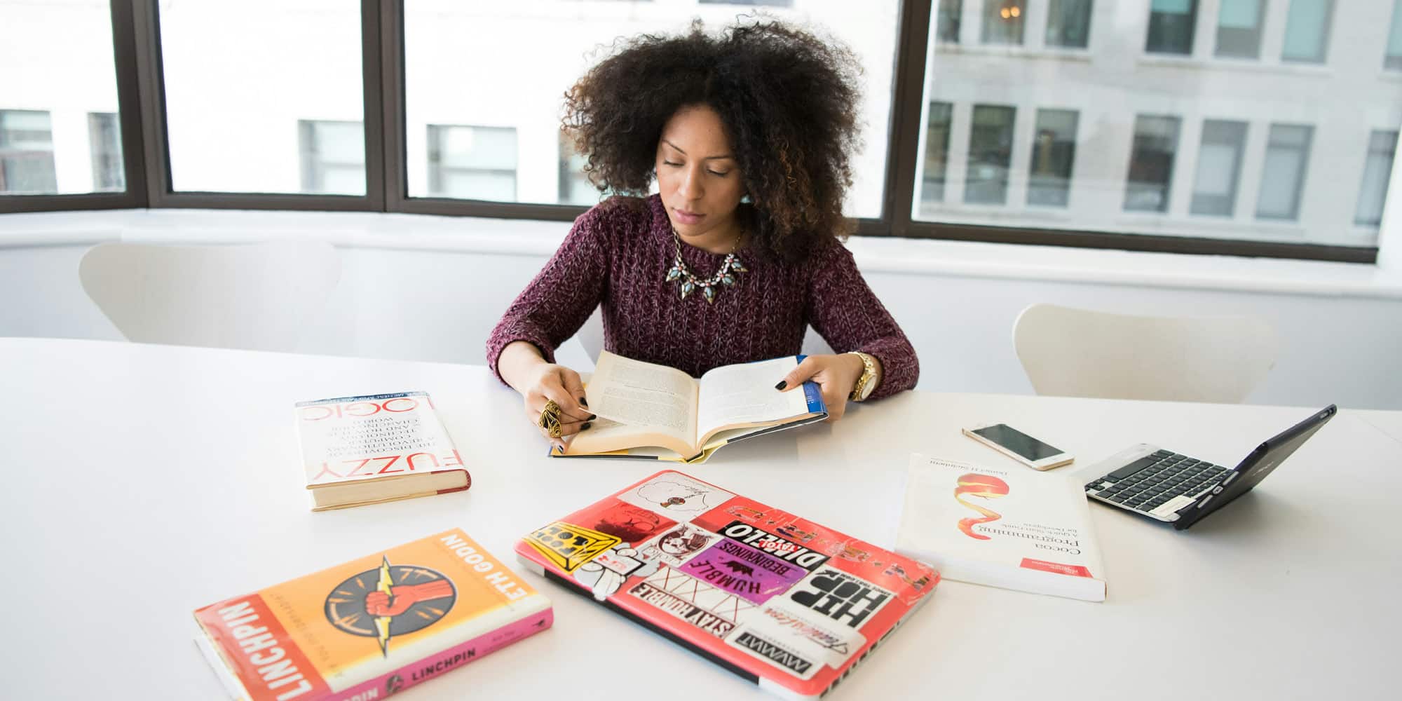 A woman with curly hair sits at a white table reading a book about the reasons why women and men are self-employed. Several books, a smartphone, and a laptop covered in stickers are on the table. Large windows reveal buildings outside.