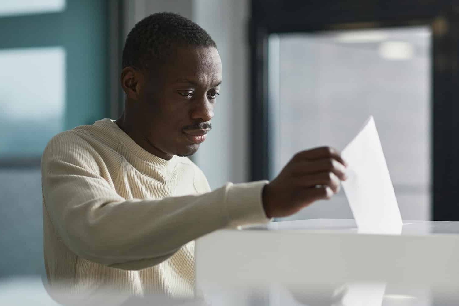 A man sits at a desk indoors, holding and examining a sheet of paper with a focused expression—perhaps pondering whether having an election that changes nothing is not such a bad outcome after all. Large windows and office furniture are visible in the background.