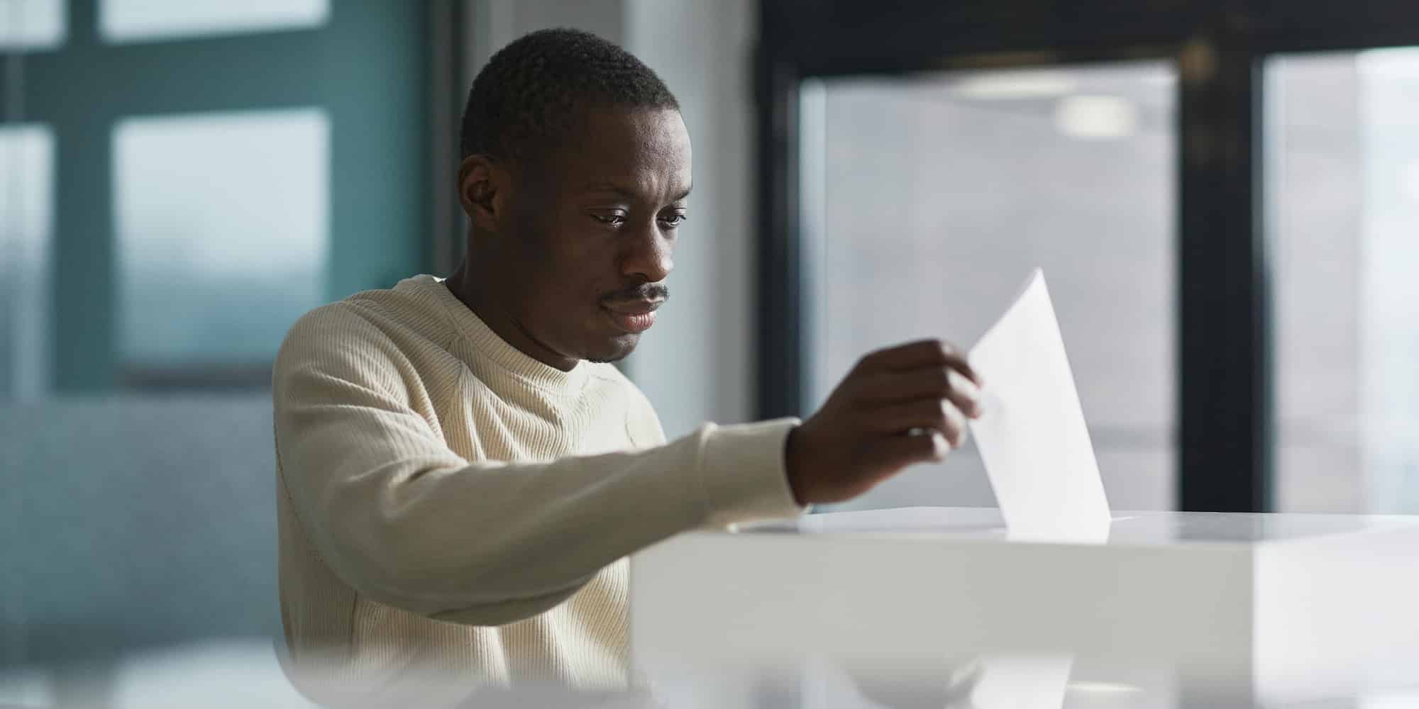 A man sits at a desk indoors, holding and examining a sheet of paper with a focused expression—perhaps pondering whether having an election that changes nothing is not such a bad outcome after all. Large windows and office furniture are visible in the background.