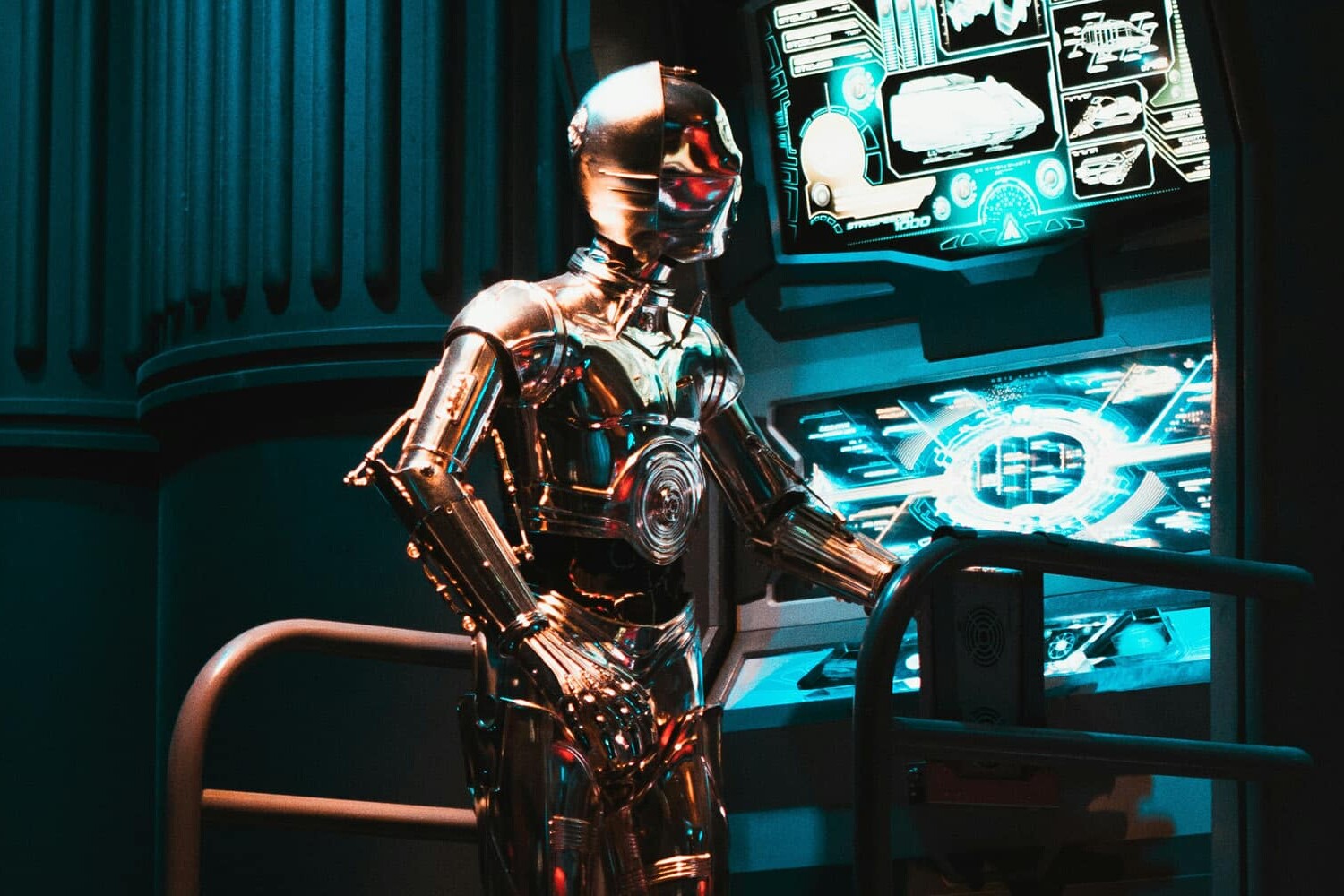 A shiny humanoid robot stands next to futuristic control panels glowing with blue light, surrounded by metallic structures in a dimly lit, sci-fi setting—raising the question: are the robots coming for our jobs?.