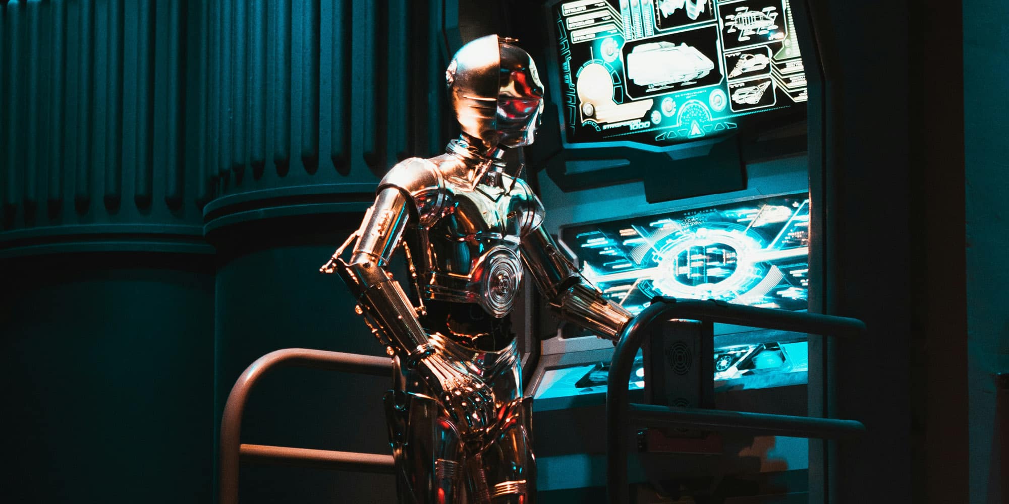 A shiny humanoid robot stands next to futuristic control panels glowing with blue light, surrounded by metallic structures in a dimly lit, sci-fi setting—raising the question: are the robots coming for our jobs?.