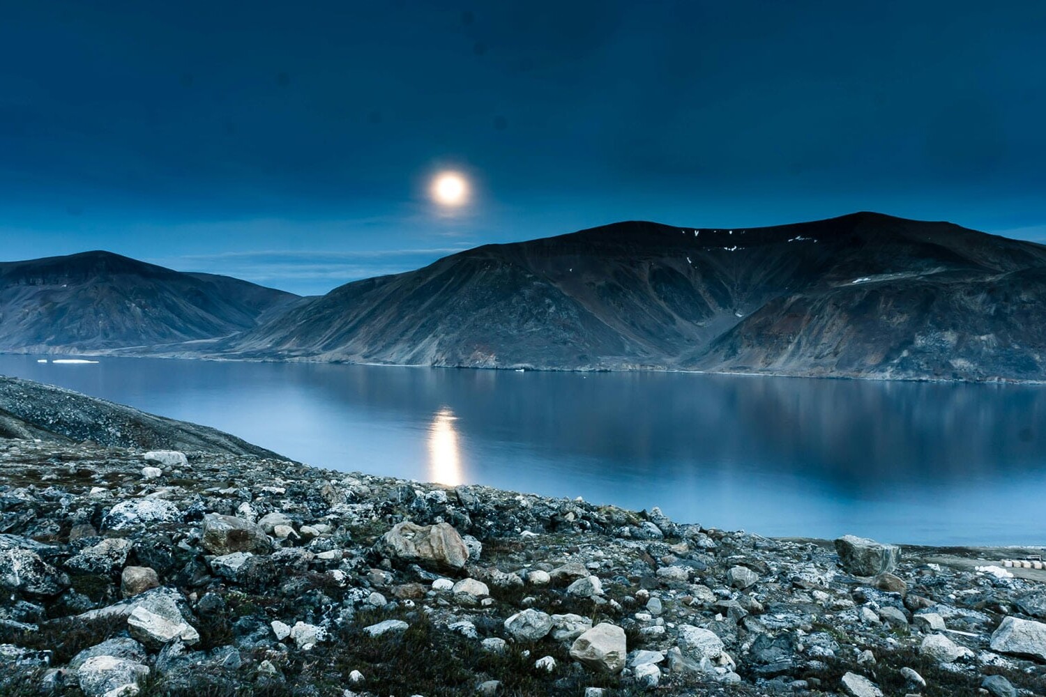 A serene arctic landscape with rocky terrain in the foreground, calm water reflecting mountains, and a bright moon illuminating the blue-toned sky—much like the shifting lens through which Canadians see the Wet’suwet’en crisis.