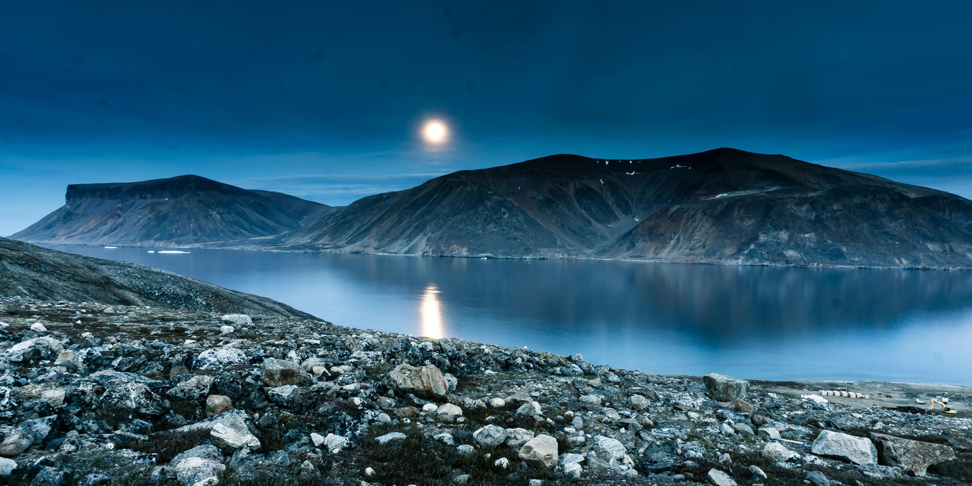 A serene arctic landscape with rocky terrain in the foreground, calm water reflecting mountains, and a bright moon illuminating the blue-toned sky—much like the shifting lens through which Canadians see the Wet’suwet’en crisis.