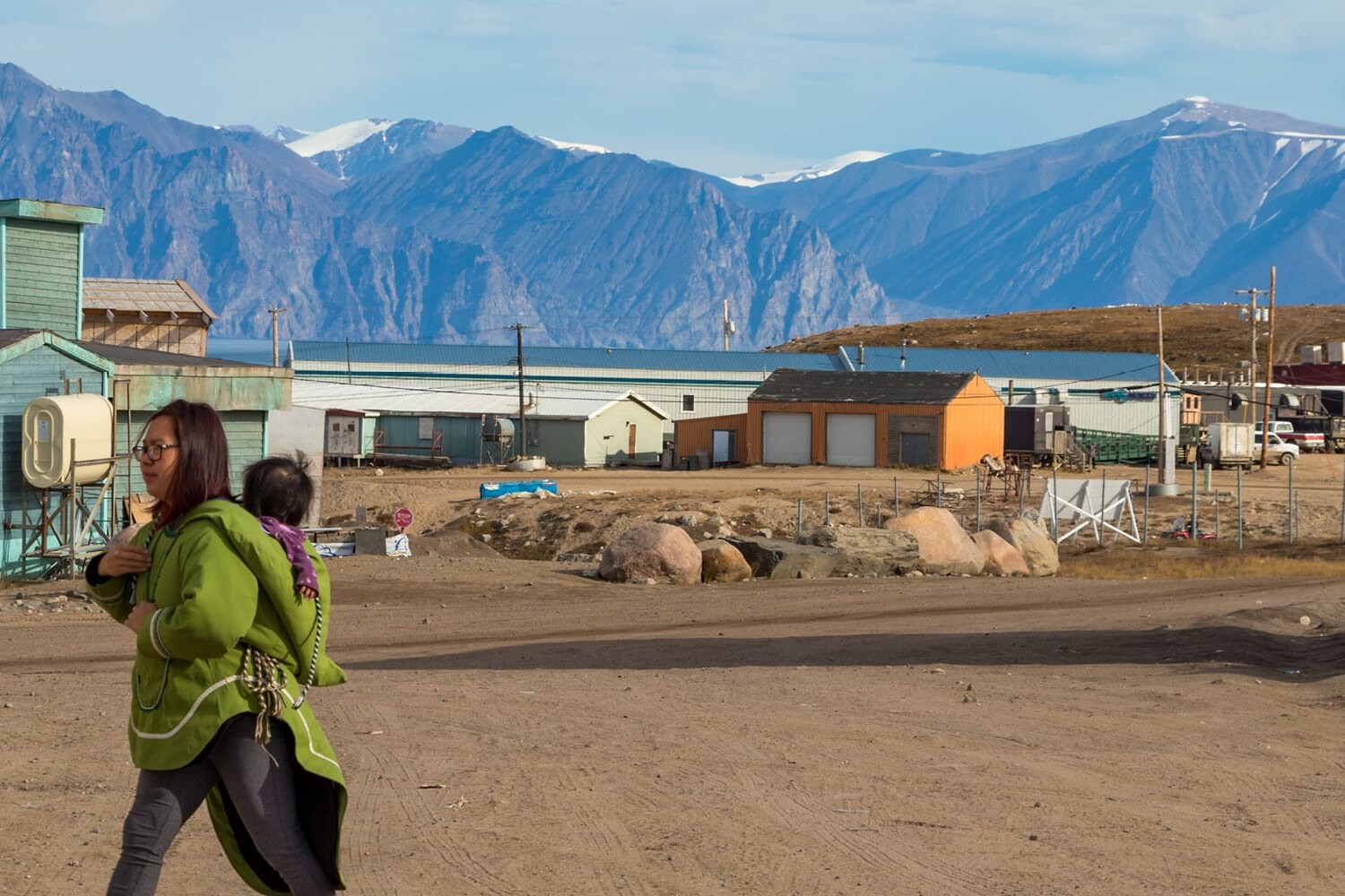 A woman in a green jacket carries a child on her back while walking through a small, remote village with colorful houses and dirt roads—echoing the shifting lens through which Canadians see the Wet’suwet’en crisis amid mountainous terrain.