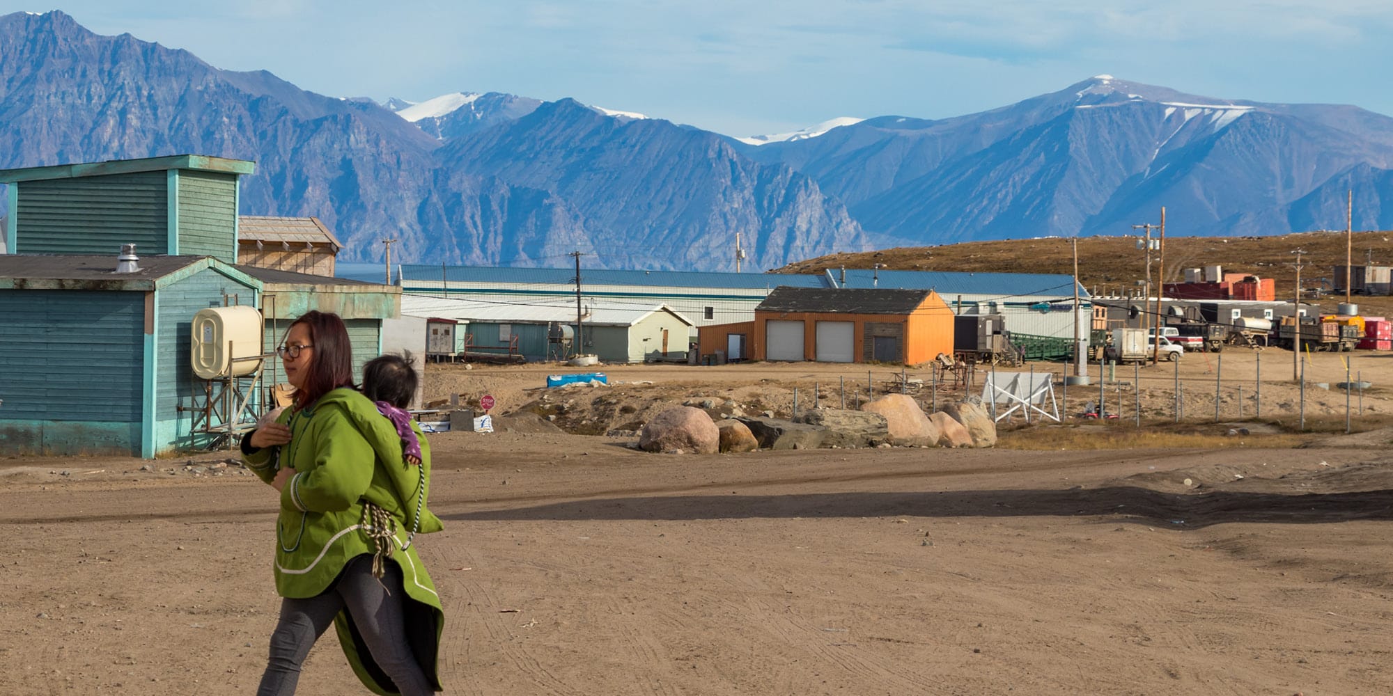 A woman in a green jacket carries a child on her back while walking through a small, remote village with colorful houses and dirt roads—echoing the shifting lens through which Canadians see the Wet’suwet’en crisis amid mountainous terrain.